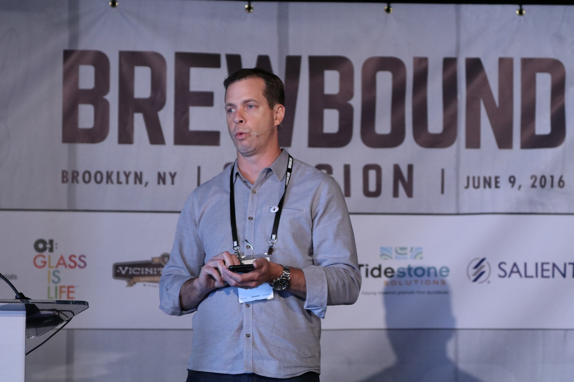 Man in a gray button-down shirt holding a phone beside a podium, in front of a Brewbound Session Brooklyn June 9, 2016 banner.