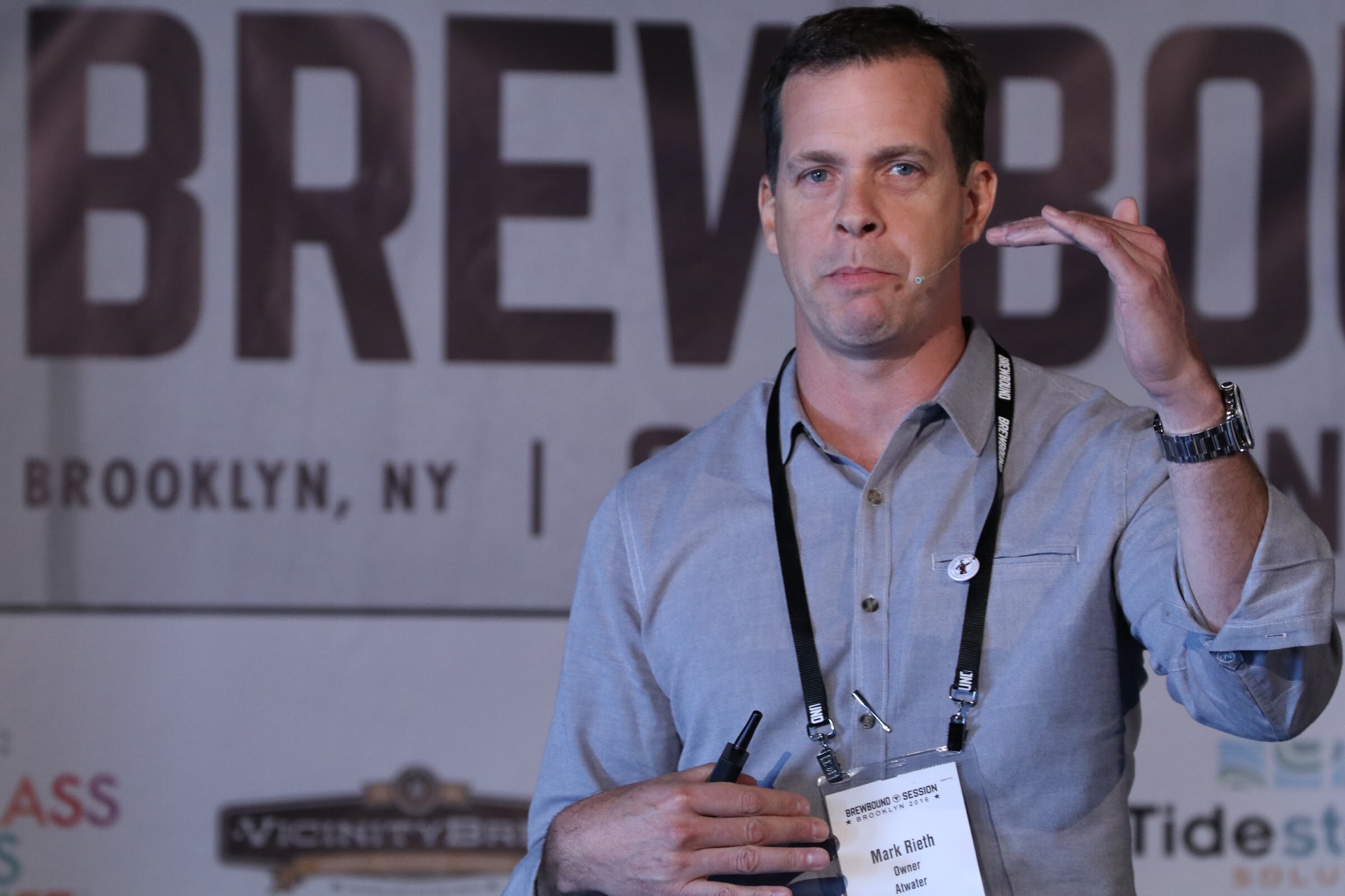 Man in a gray shirt with a speaker badge gesturing with one hand and holding a clicker in front of the Brewbound banner.
