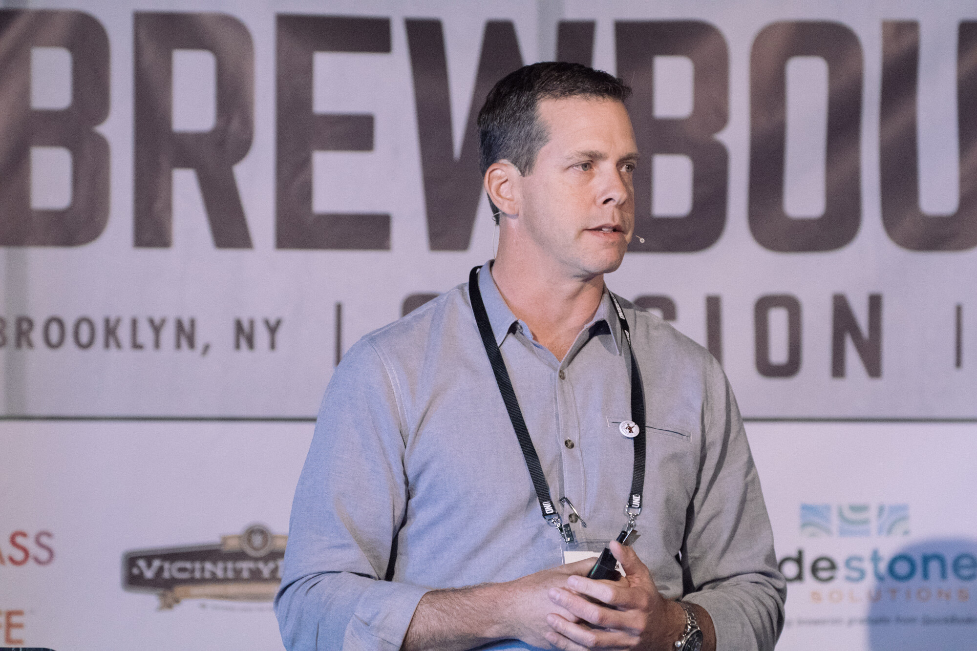 Man in a gray shirt with a lanyard holding a presentation clicker in front of a Brewbound Session Brooklyn banner.