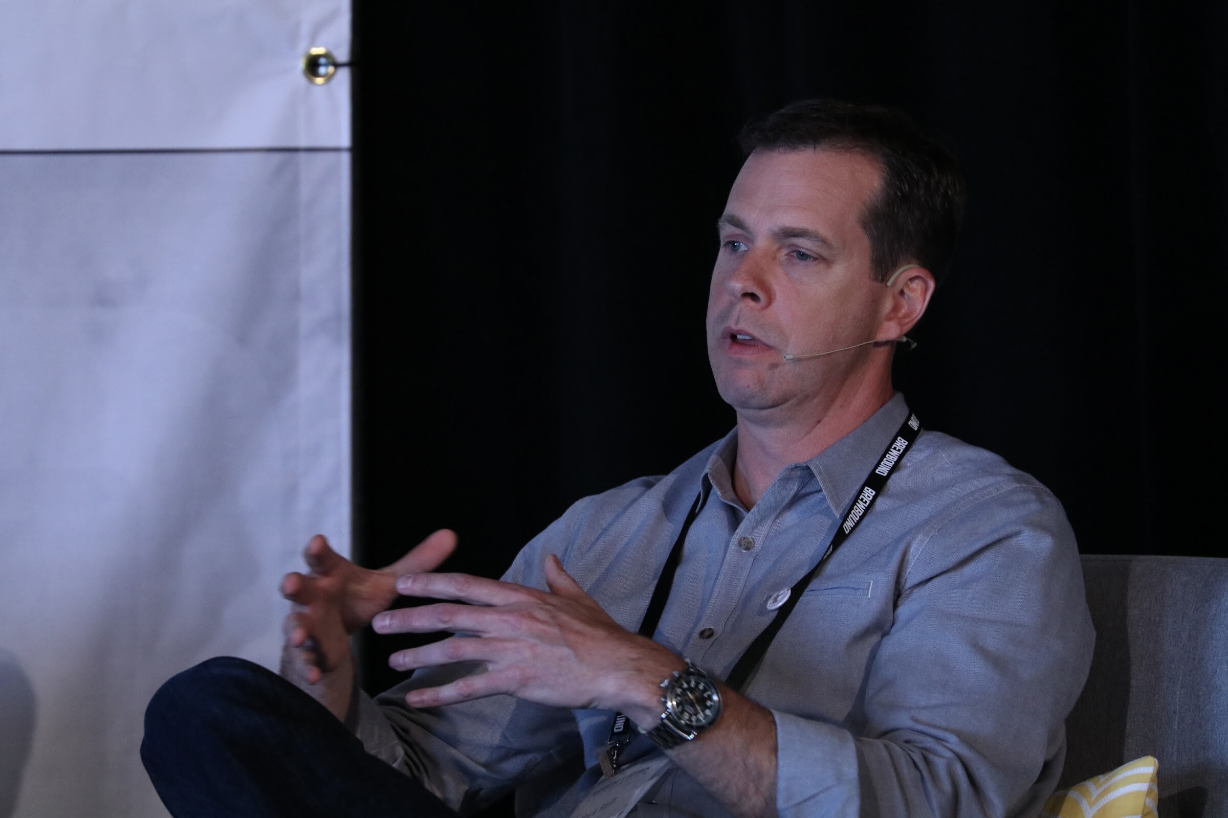 Man in a gray shirt with a headset microphone seated and gesturing with both hands against a black curtain.