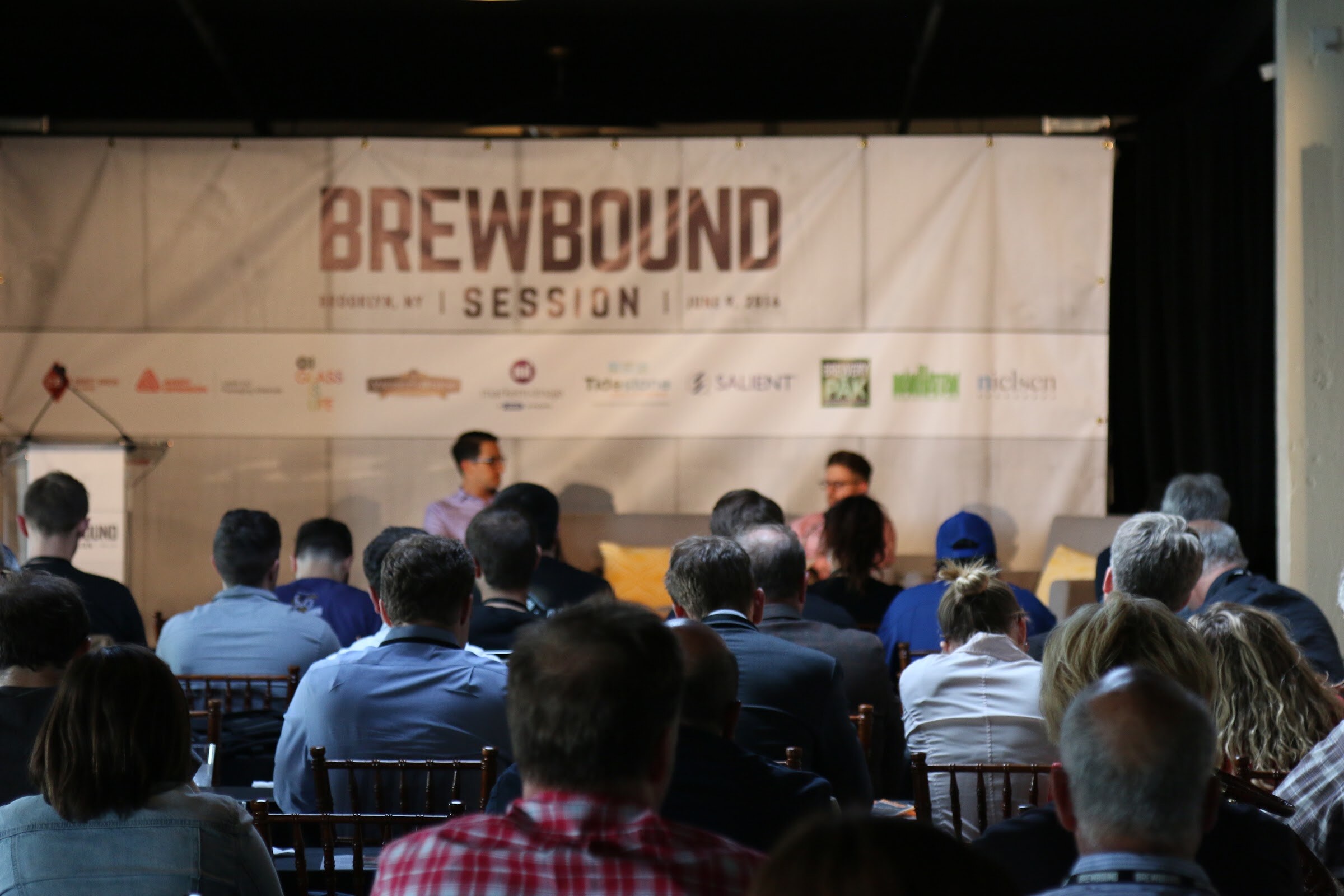 Rows of audience members from behind facing a stage with two people on couches under a Brewbound Session banner with sponsor logos.