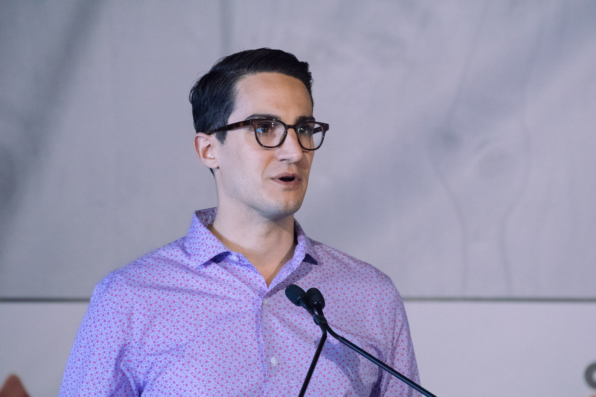 Dark-haired man with glasses in a purple patterned shirt speaking at a lectern with two microphones.