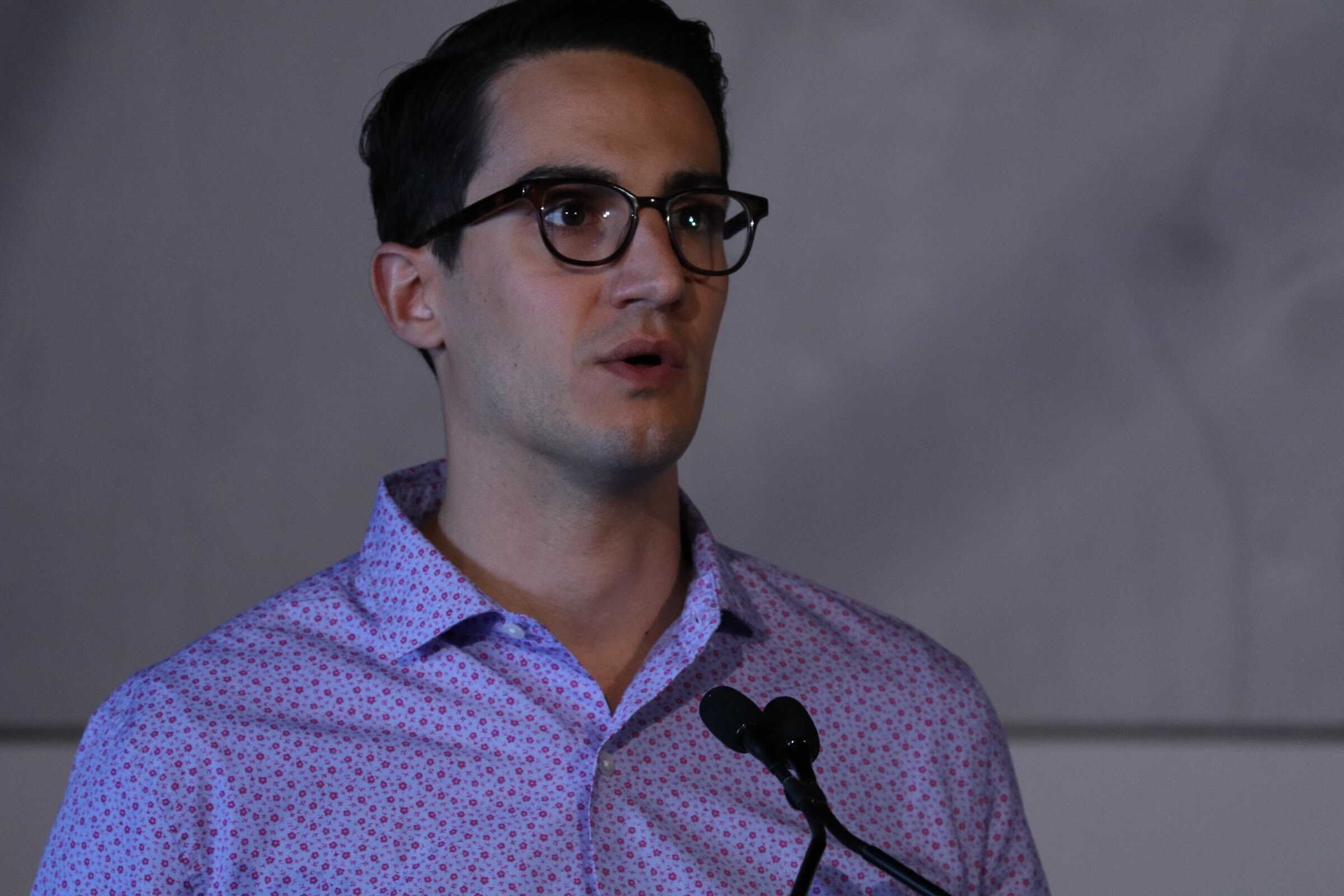 Close view of a dark-haired man in glasses and a purple patterned shirt speaking at a lectern.