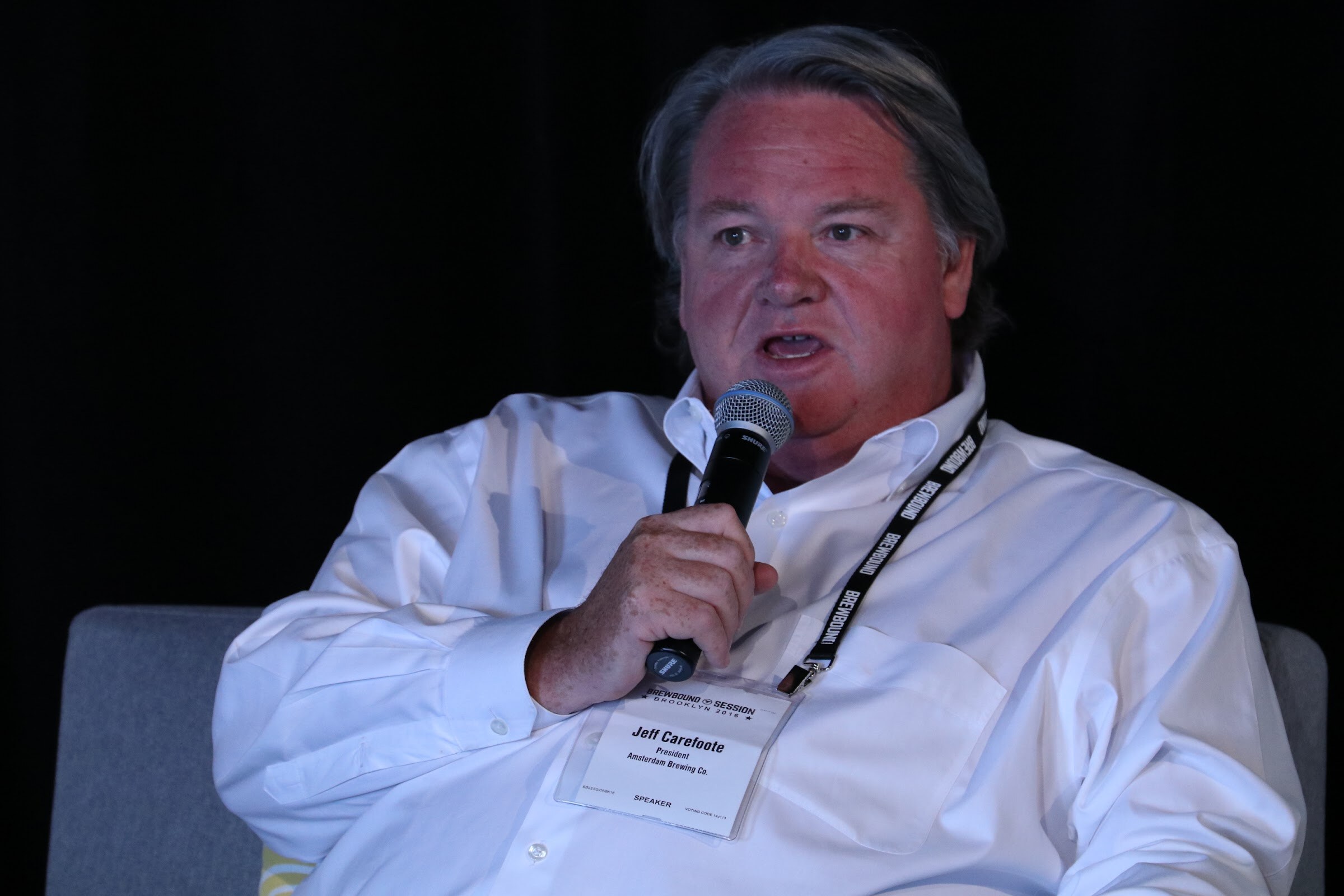 Man in a white shirt with a speaker badge holding a microphone against a black curtain.