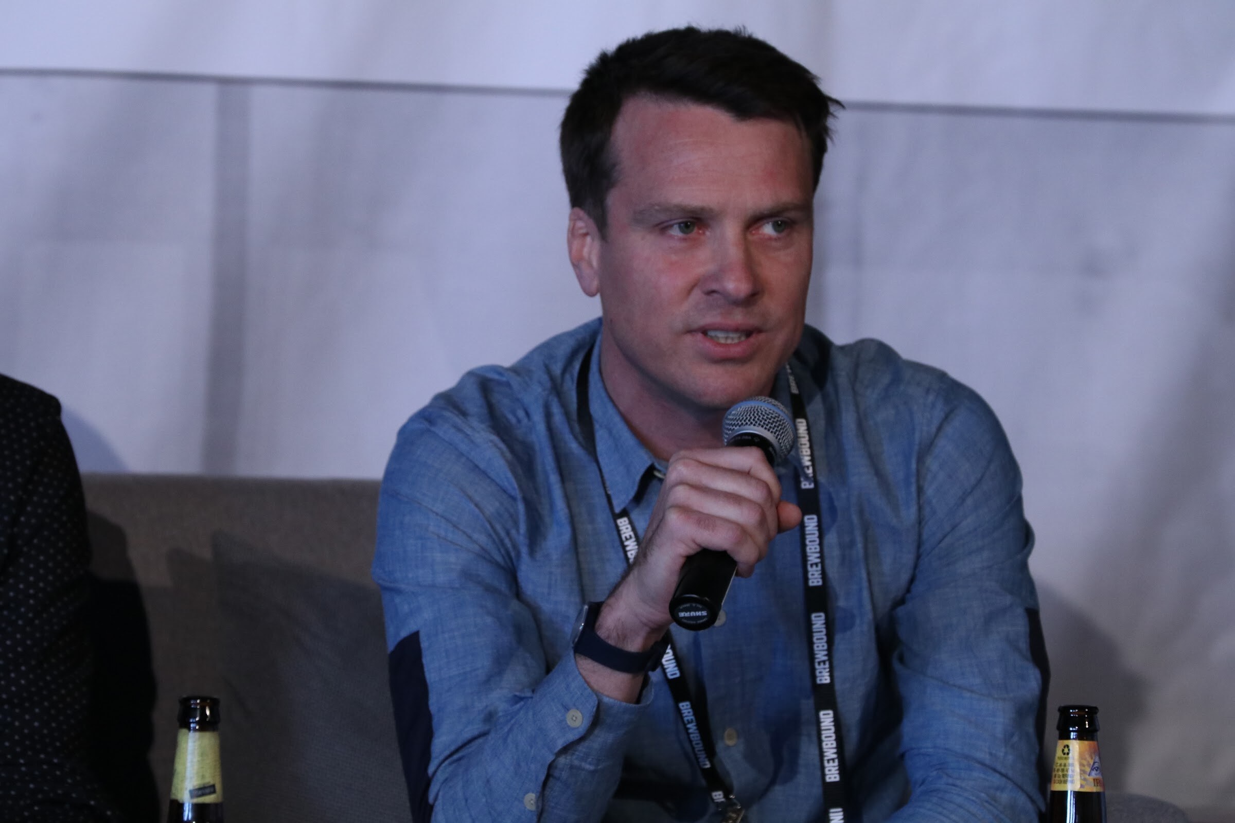 Man in a blue shirt holding a microphone, seated on a couch with two beer bottles at the edge of the frame.