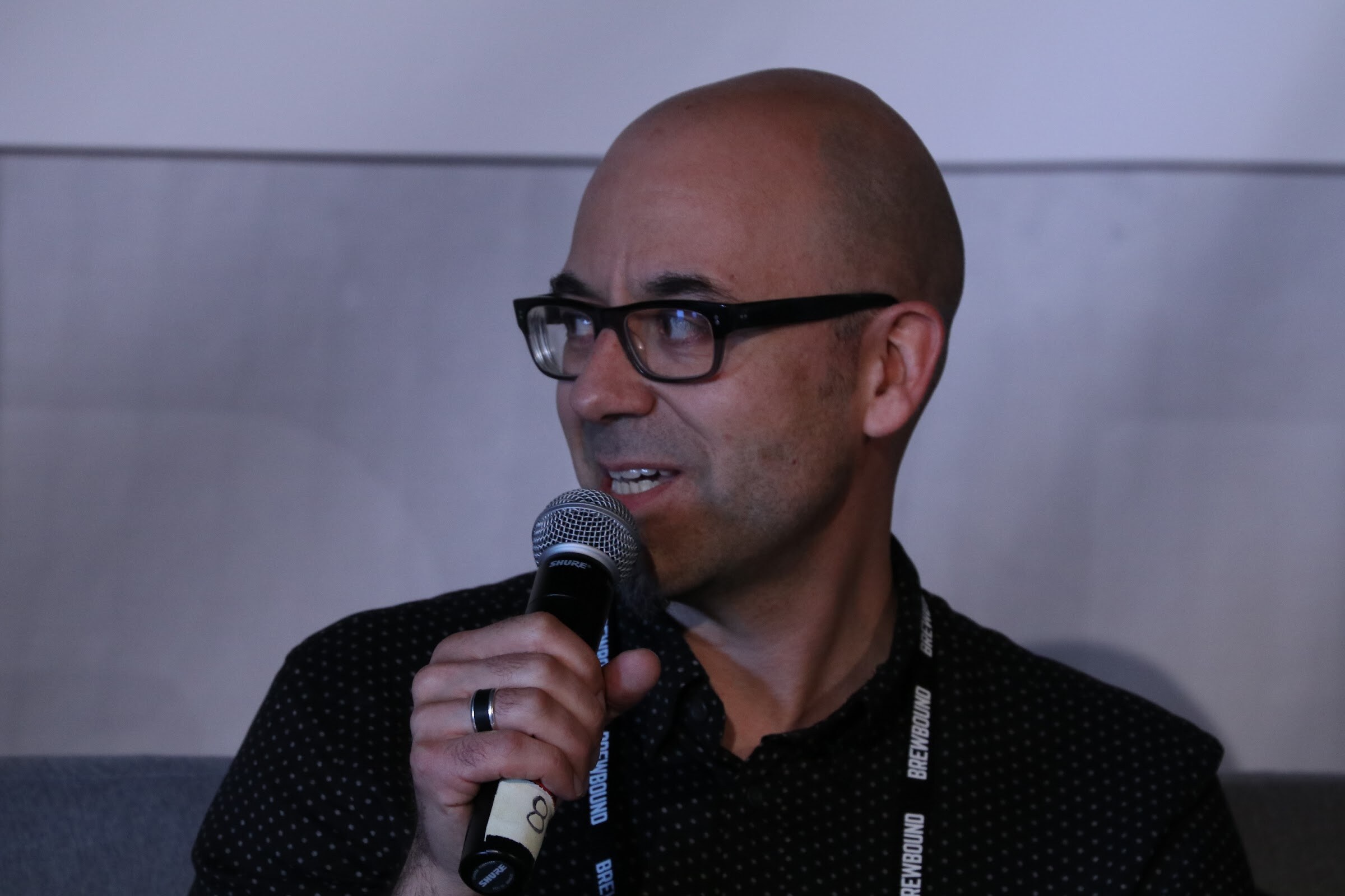 Bald man with dark-rimmed glasses holding a microphone and looking to his left.
