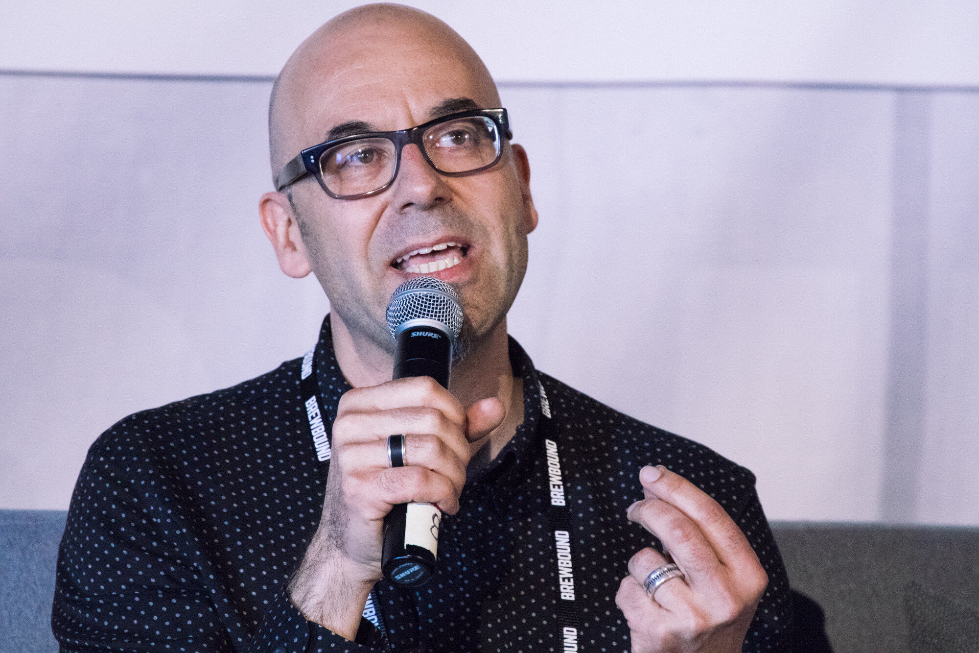 Bald man in glasses and a dark dotted shirt holding a microphone and raising one hand.