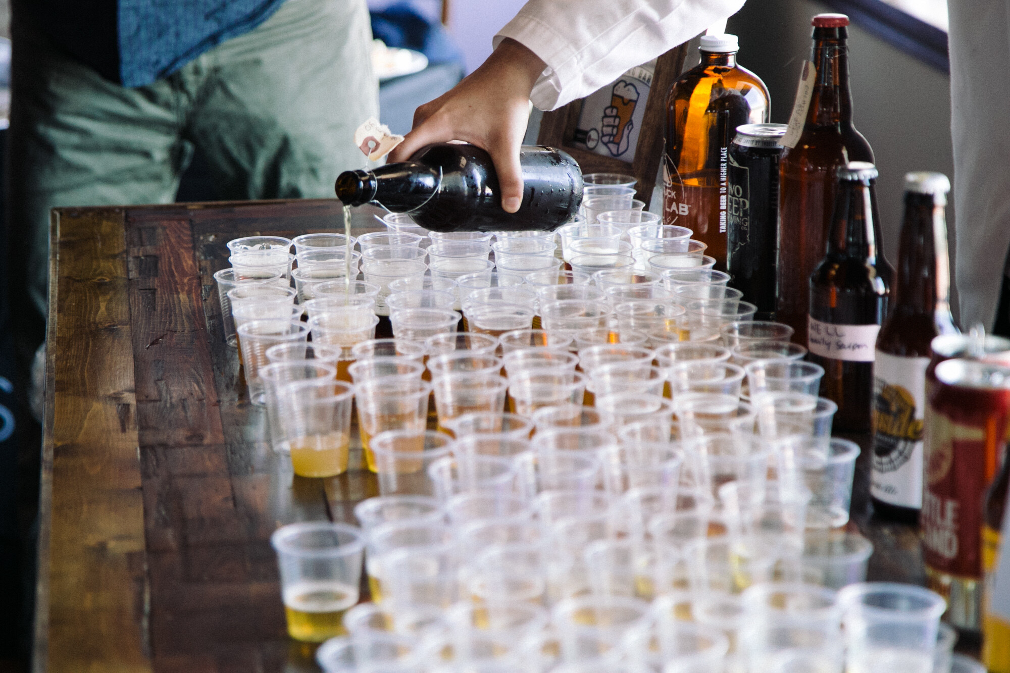 A hand pouring beer from a dark bottle into dozens of clear plastic cups on a wooden table, with growlers and cans at the right.