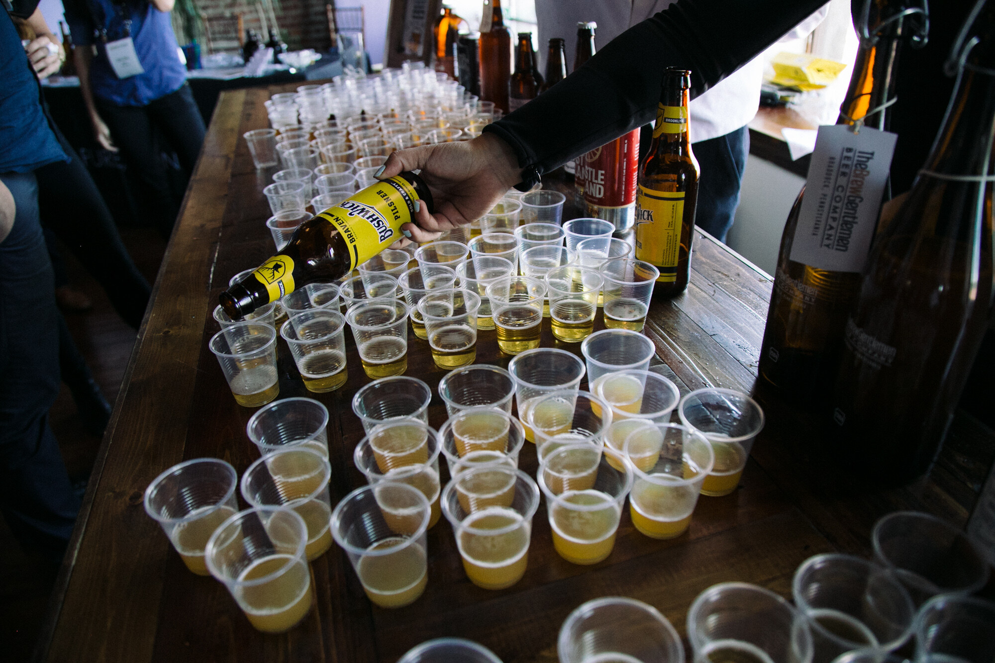 Hand pouring from a yellow-labeled Bushwhacker Pilsner bottle into plastic cups on a long wooden table.