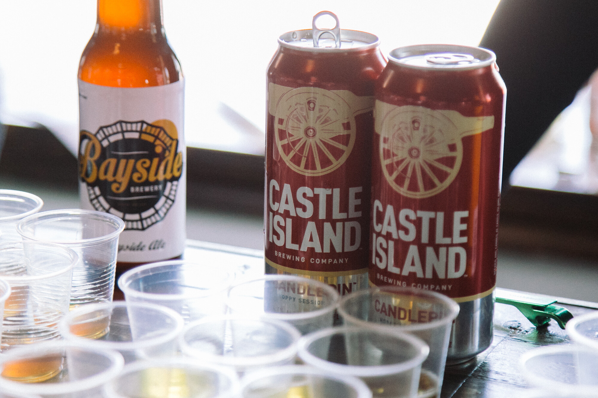 Bottle labeled Bayside Brewery next to two red Castle Island Brewing Company cans and clear plastic cups.