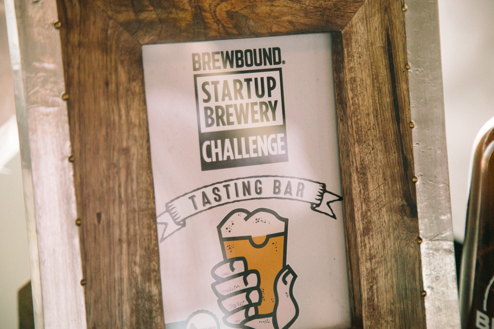 Wood-framed sign reading Brewbound Startup Brewery Challenge Tasting Bar with an illustration of a hand holding a beer.
