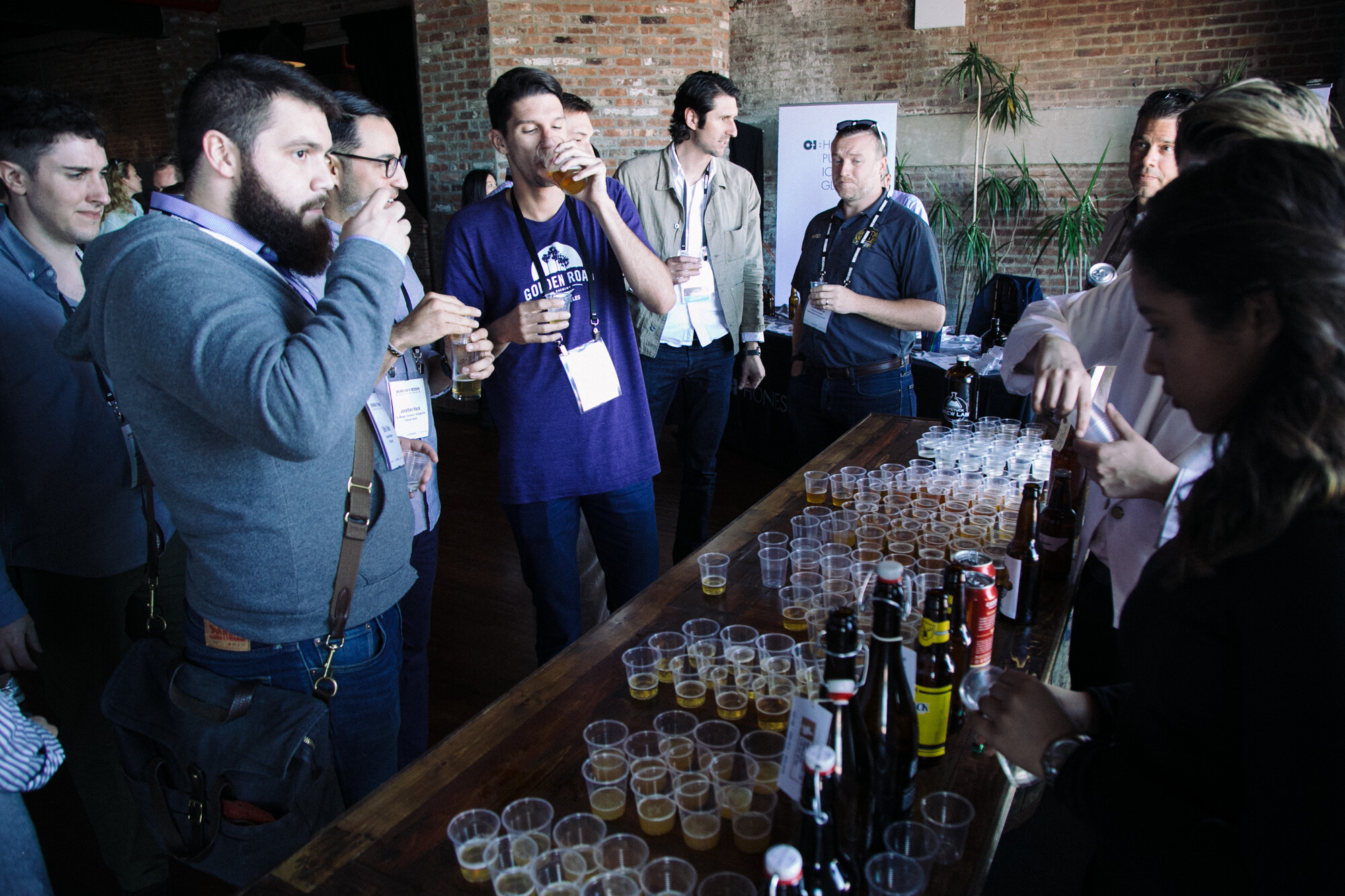 Group of attendees with lanyards drinking from small cups around a long wooden table of sample cups, in a room with brick walls.