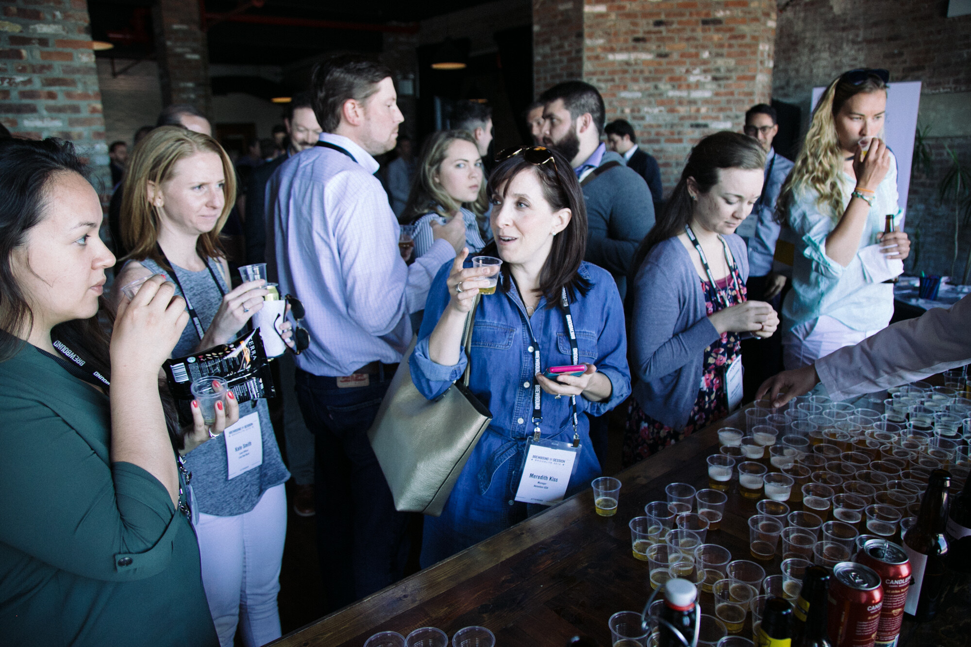 Several women with lanyards holding small sample cups beside a long table of plastic cups and cans.