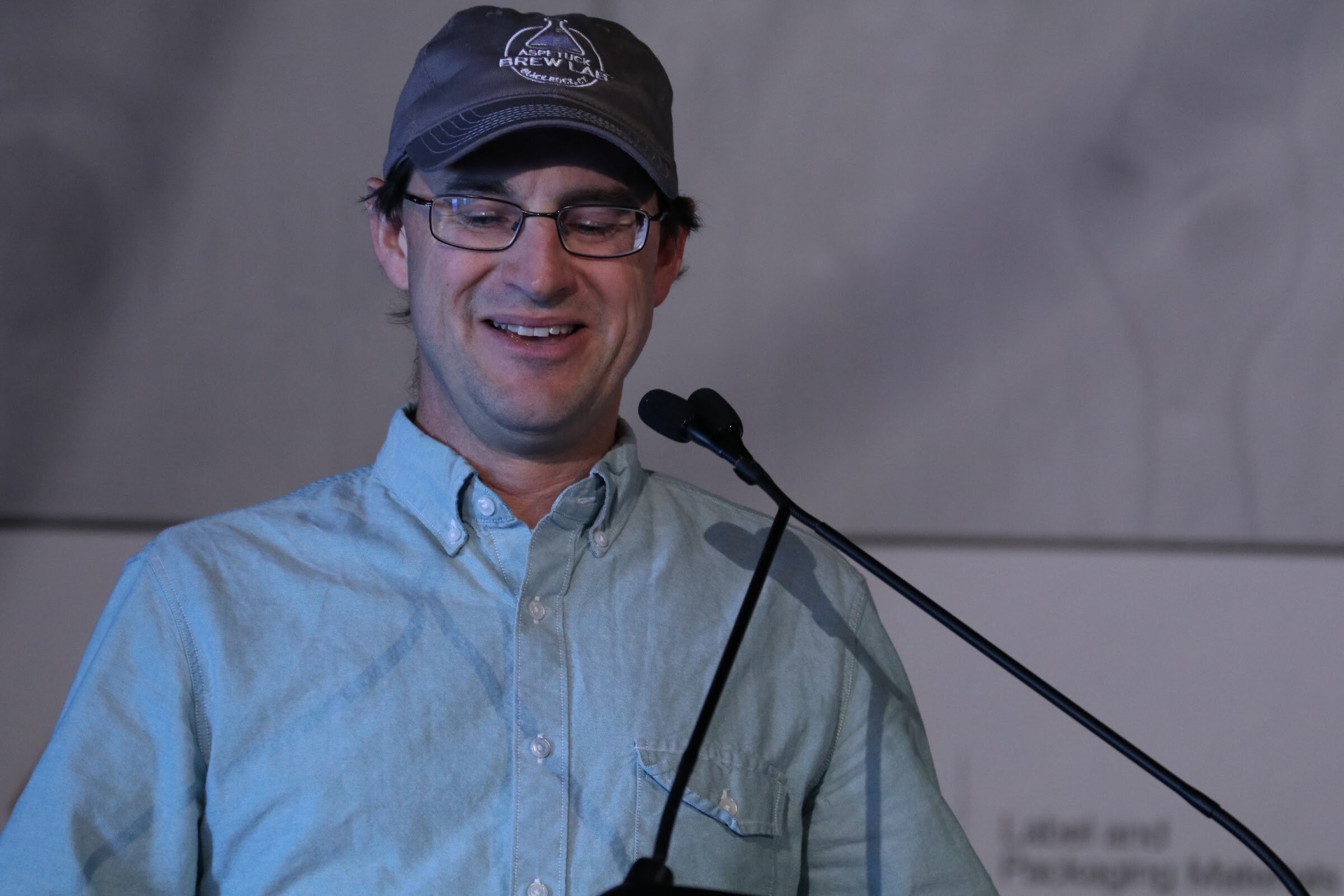 Man in a gray baseball cap and glasses wearing a light blue shirt smiling at a lectern with two microphones.
