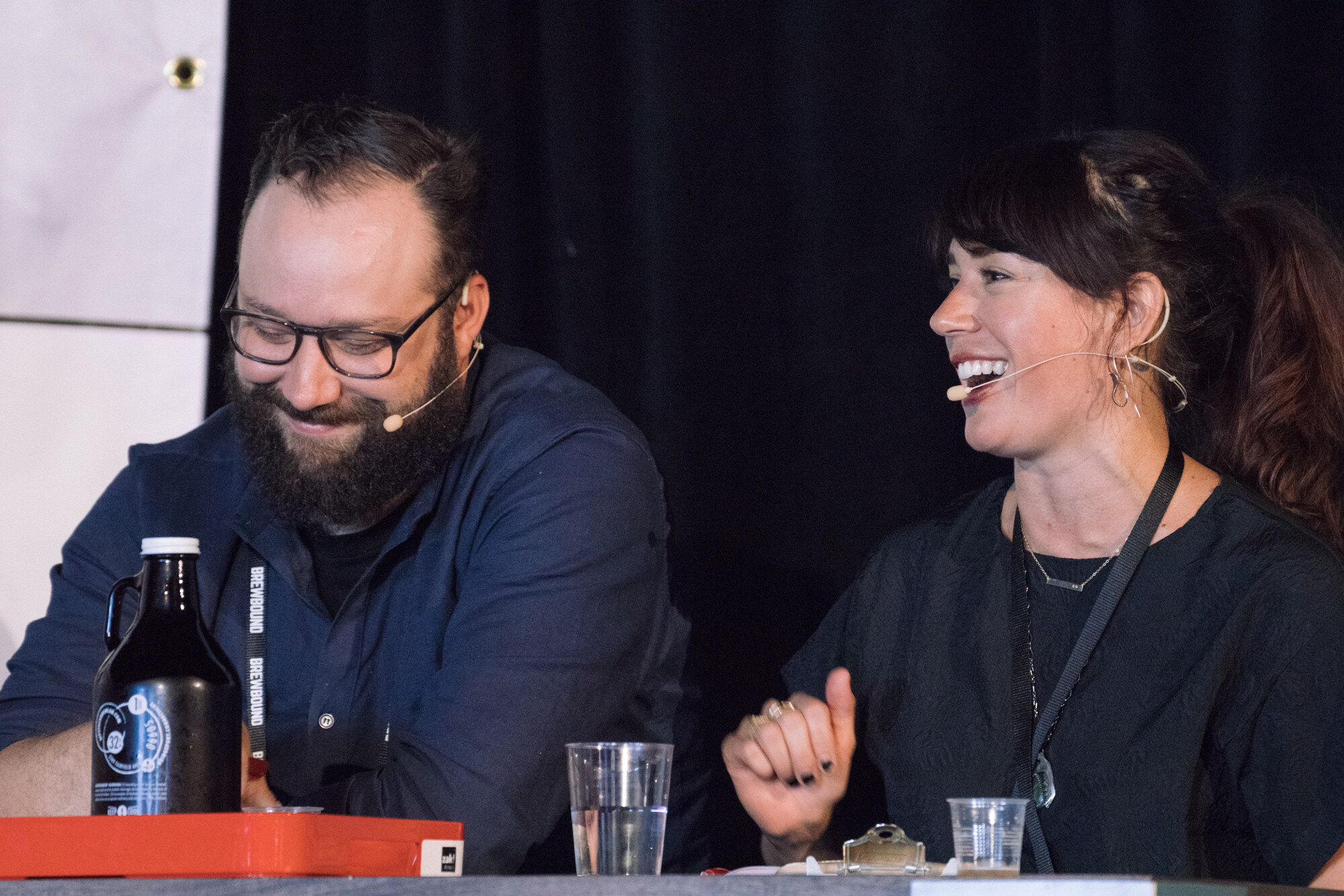 Bearded man in glasses looking down and a laughing woman with a headset microphone at a table with a black growler and plastic cups.