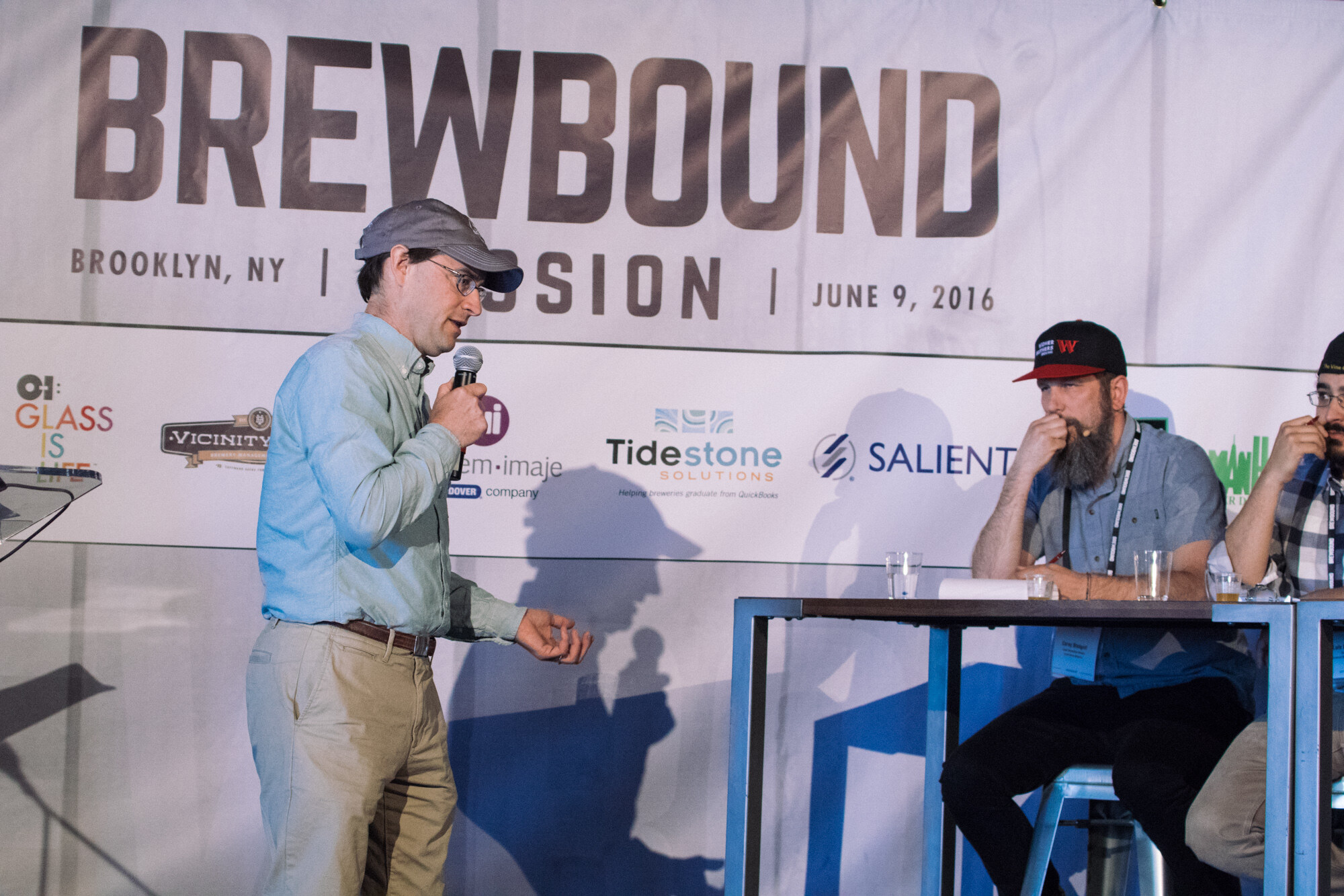 Man in a light blue shirt and gray cap holding a microphone on stage as two seated judges look on, under the Brewbound banner.