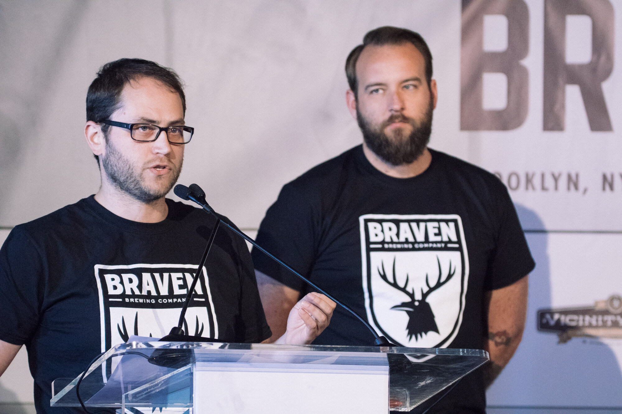 Two men in black Braven Brewing Company t-shirts at a clear acrylic lectern, one speaking into a microphone.