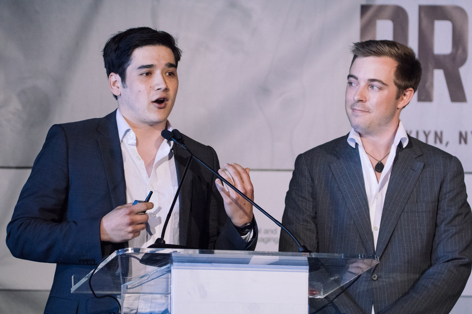 Two men in suit jackets at a lectern, one speaking and gesturing with a clicker while the other looks on.