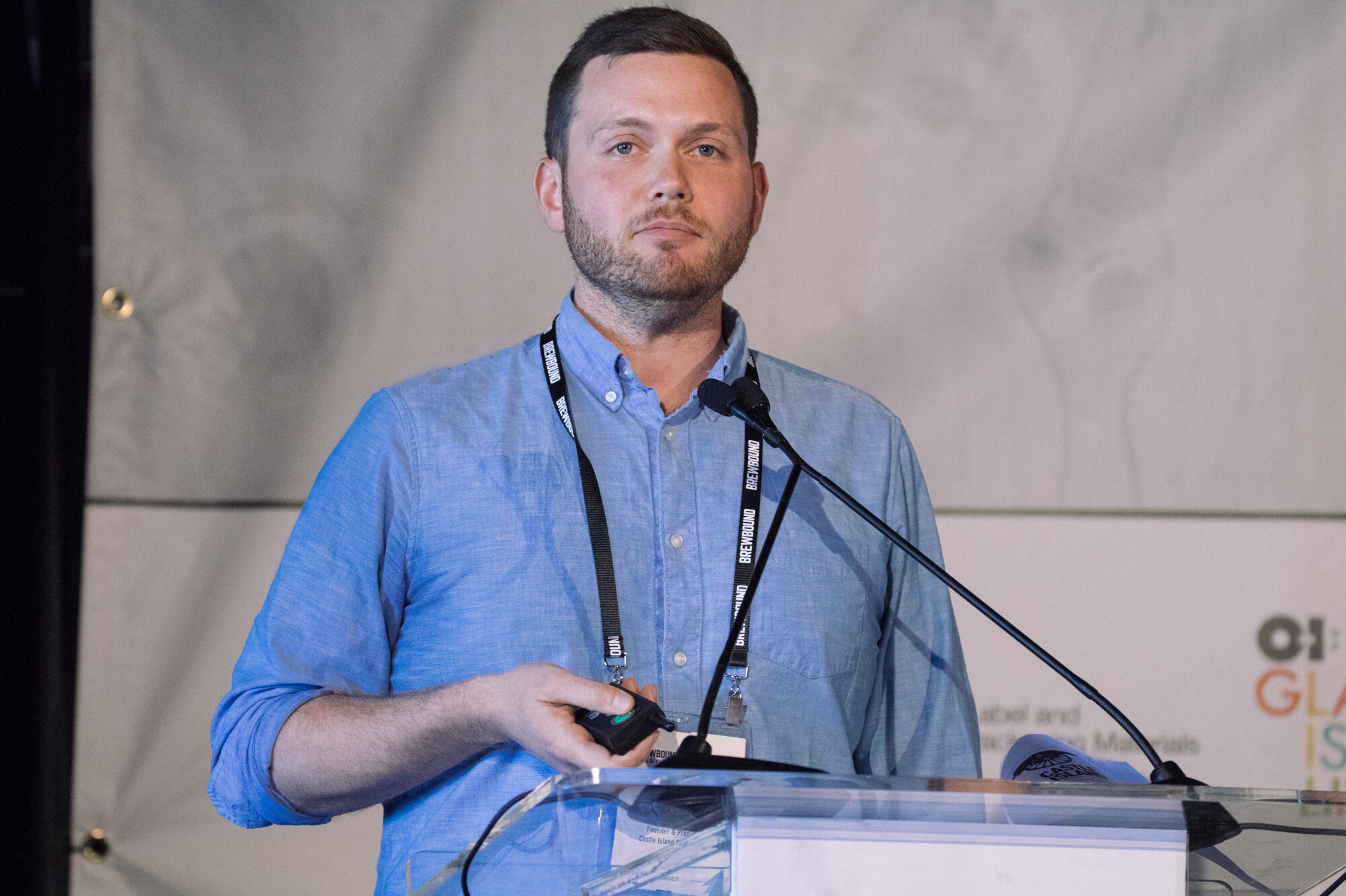 Bearded man in a blue shirt with a Brewbound lanyard holding a presentation clicker at a lectern.