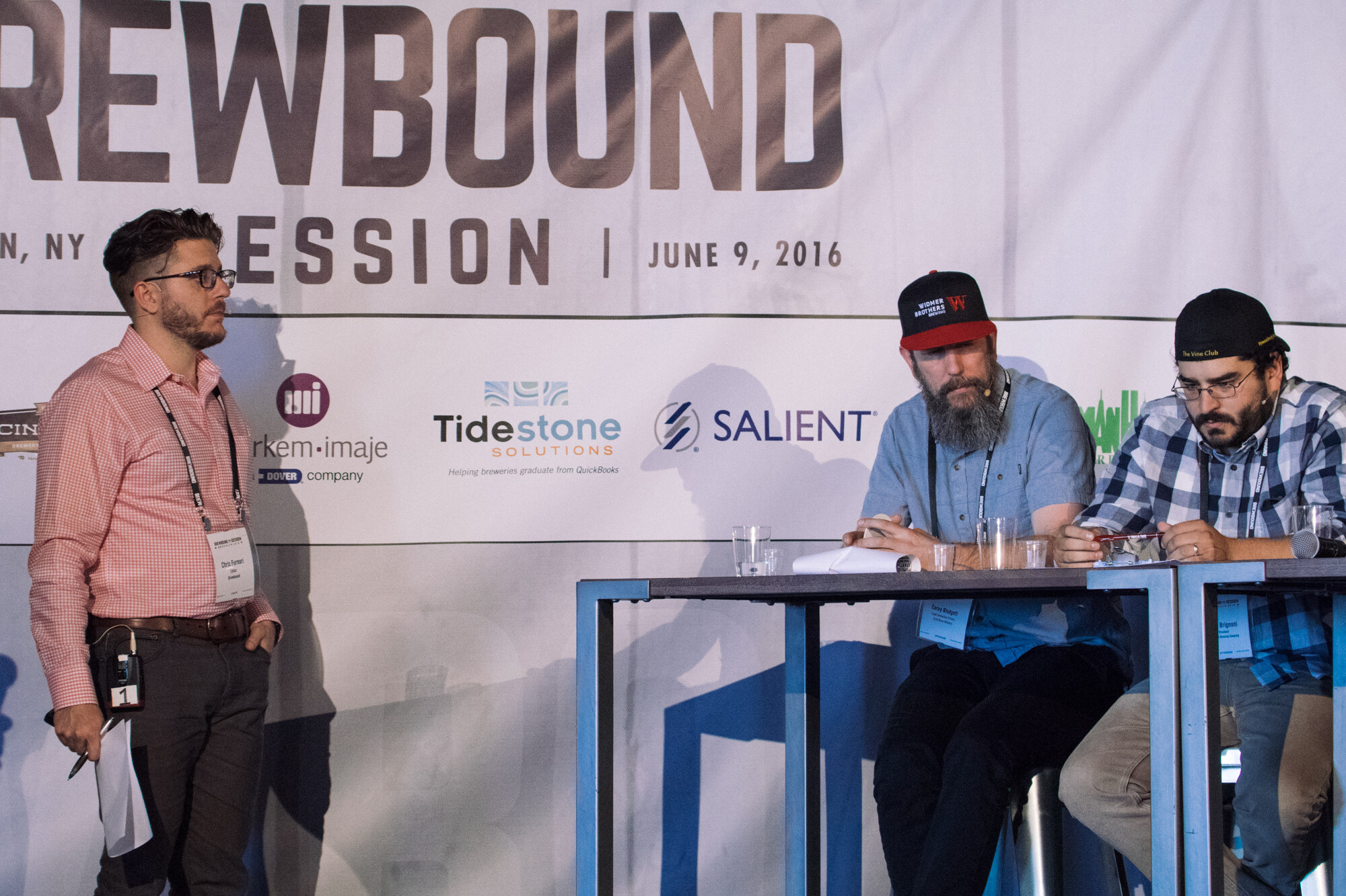 Man in a pink checked shirt holding papers beside two seated judges at a high table under a Brewbound Session banner dated June 9, 2016.