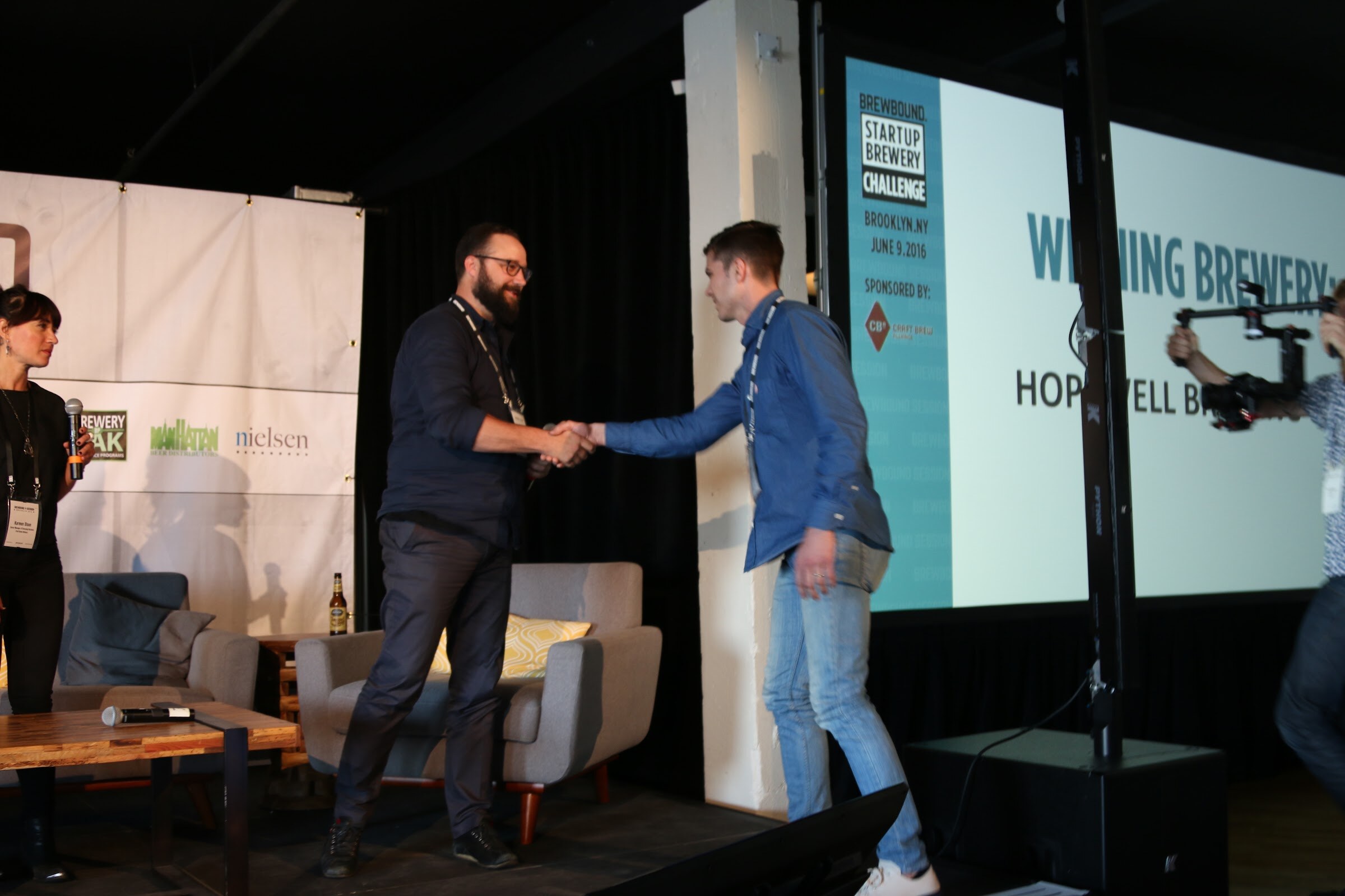 Two men shaking hands in front of a screen reading Brewbound Startup Brewery Challenge, Winning Brewery: Hopwell Brewing Co., with a camera operator at the right.