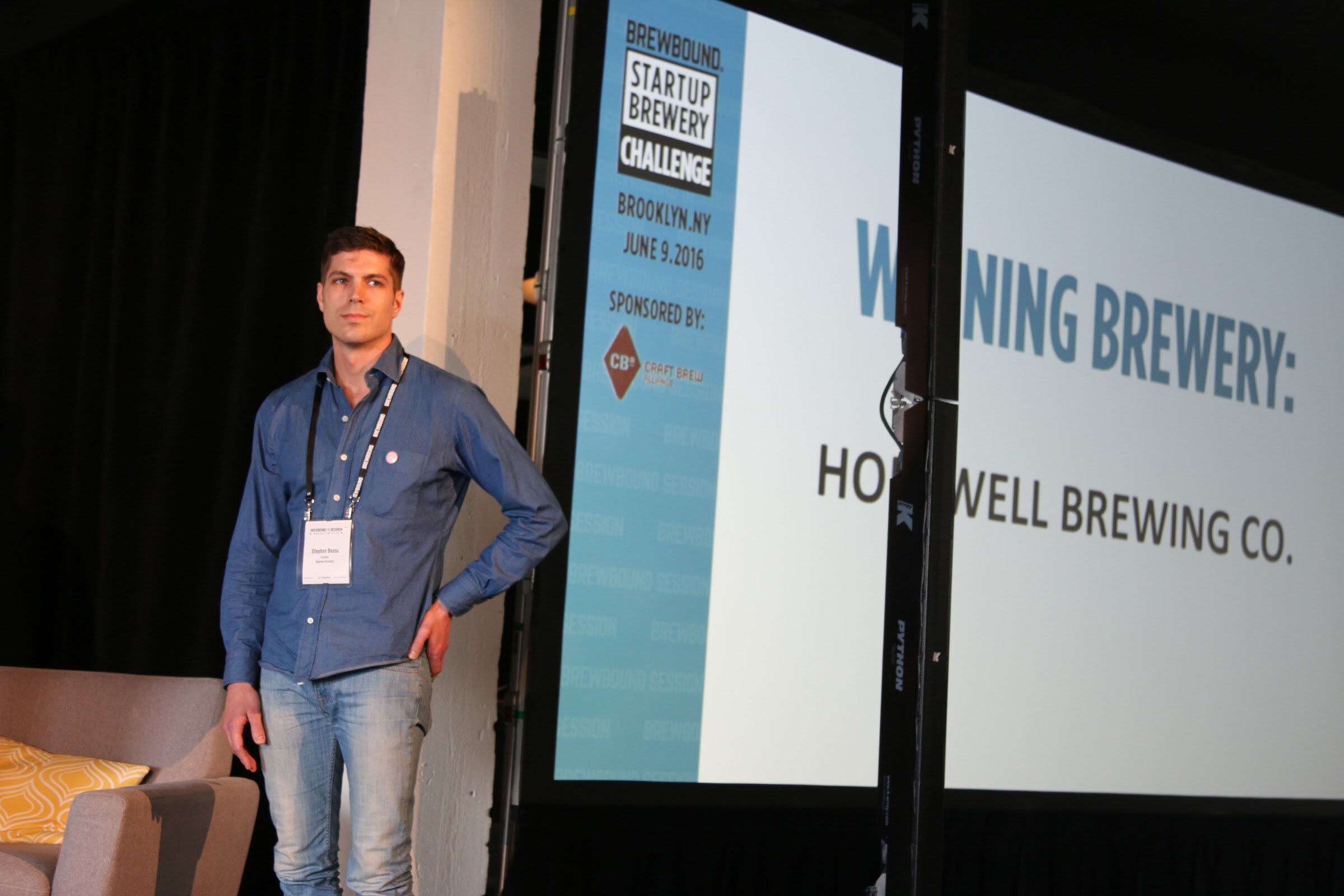 Man in a blue button-down shirt standing with a hand on his hip beside a screen reading Winning Brewery: Hopwell Brewing Co. in the Brewbound Startup Brewery Challenge graphic.