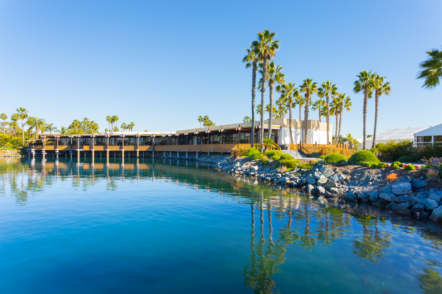 Low wooden waterfront building with palm trees along a rocky shore, reflected in still blue water under a clear sky.