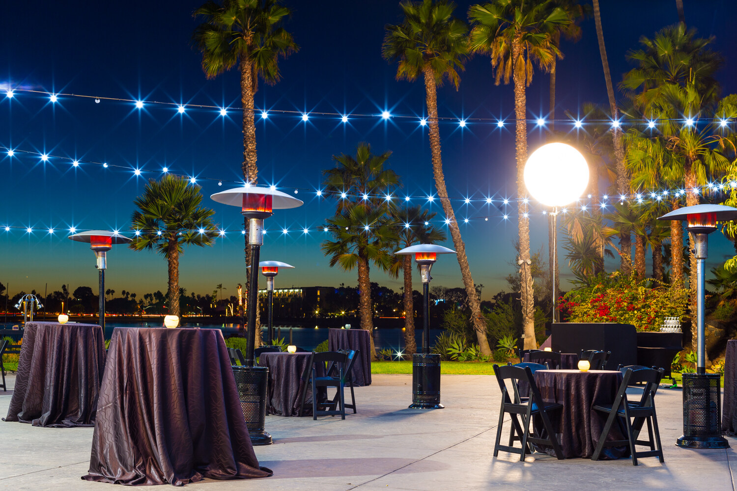 Empty patio at dusk with draped high-top tables, glowing patio heaters, string lights, palm trees and a lit globe lamp, with the water and city lights behind.