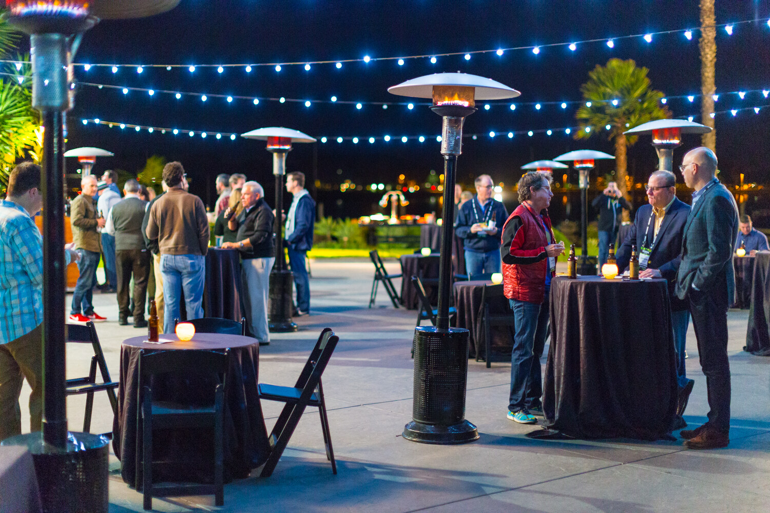 Evening patio scene with groups talking at draped high-top tables beside glowing patio heaters, string lights overhead and city lights across the water.