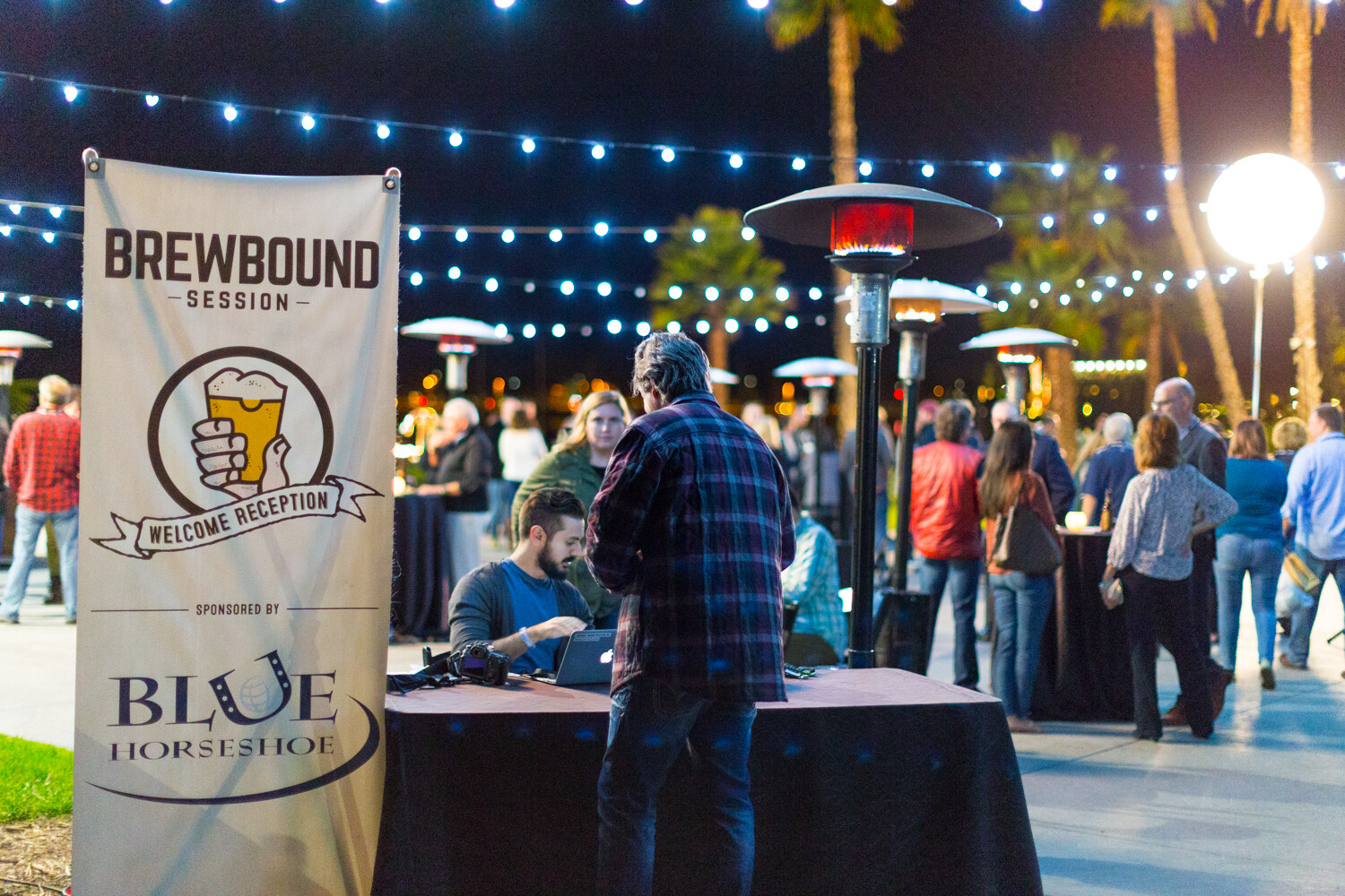 Tall banner reading Brewbound Session Welcome Reception sponsored by Blue Horseshoe beside a table where a man works on a laptop and another attendee leans over it, with a crowd and string lights behind.