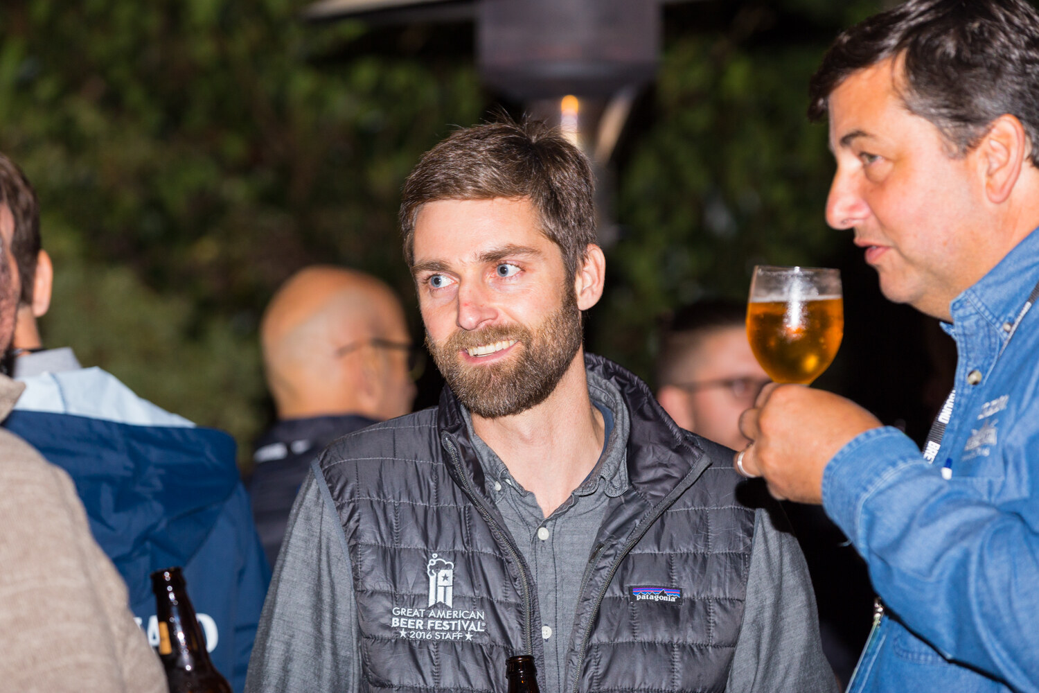 Man in a gray puffer vest with a Great American Beer Festival 2016 Staff logo looking off-camera while a man in a denim shirt holds up a glass of beer beside him, outdoors at night.