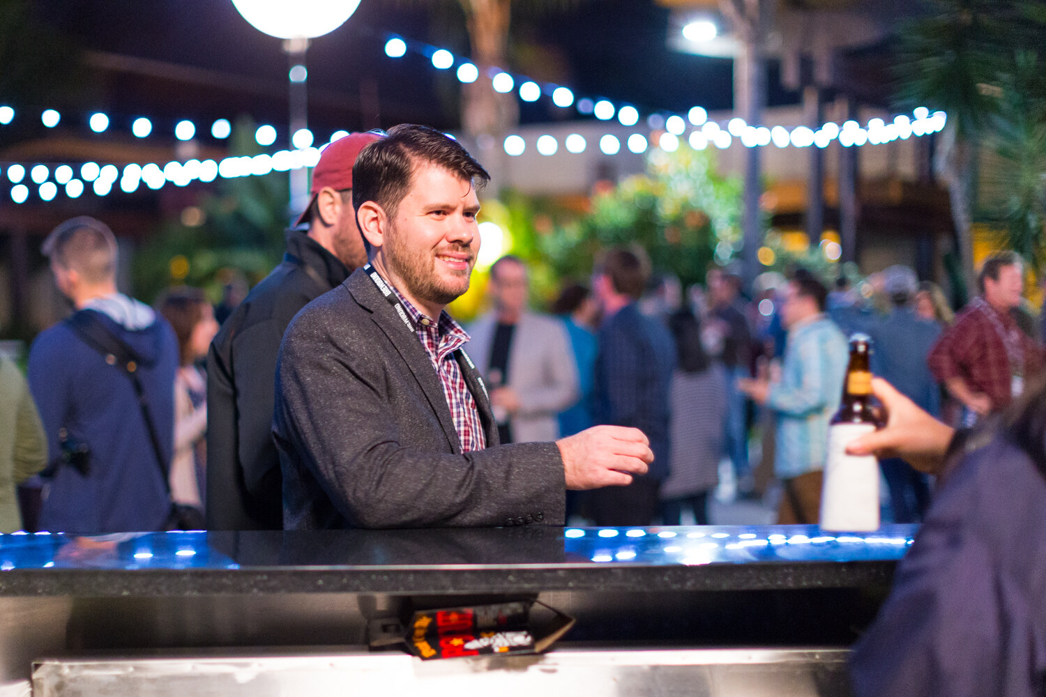 Bearded man in a gray blazer and plaid shirt with a lanyard resting an arm on a bar top, with blurred string lights and guests behind and a hand holding a beer bottle at right.