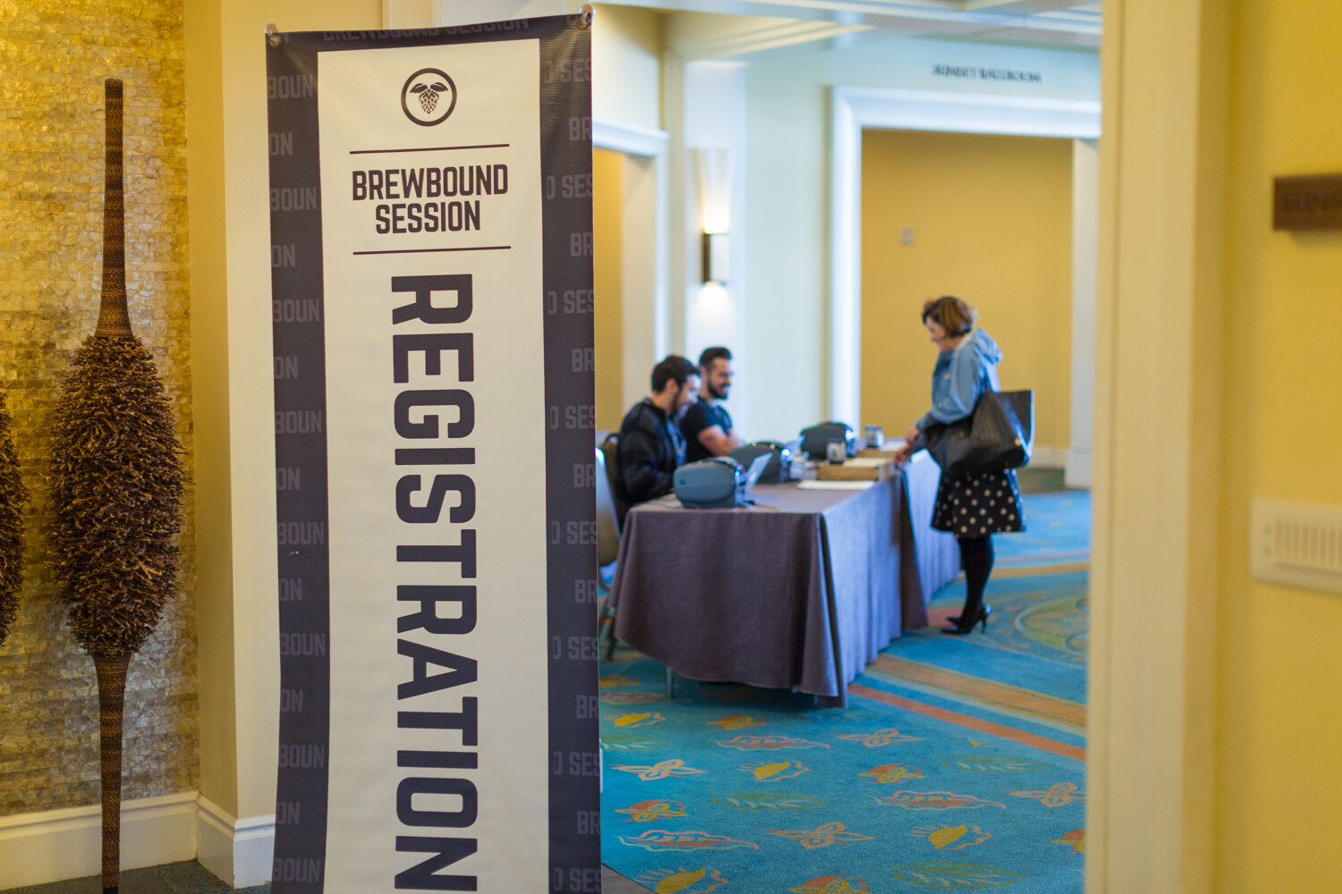 Vertical banner reading Brewbound Session Registration beside a draped table with two staff members and a woman in a polka-dot skirt standing at it, on a patterned blue carpet.