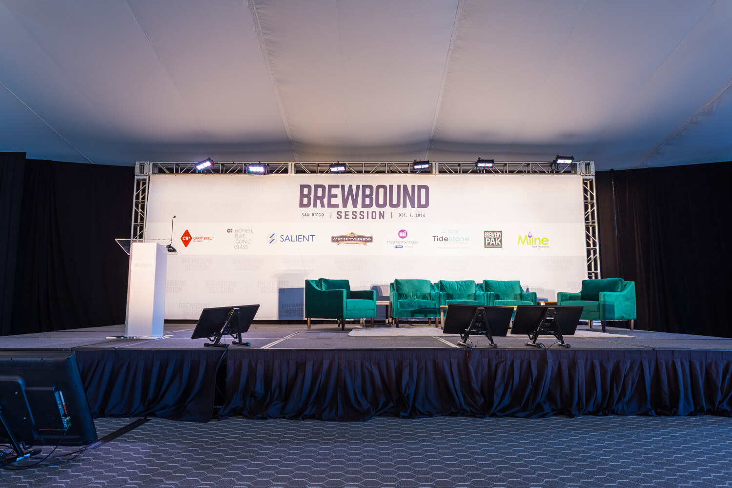 Empty stage with a white lectern and teal green velvet armchairs in front of a banner listing sponsors including Craft Brew Alliance, Salient, Vicinity Brew, Markem-Imaje, Tidestone, Brewery Pak and Milne.