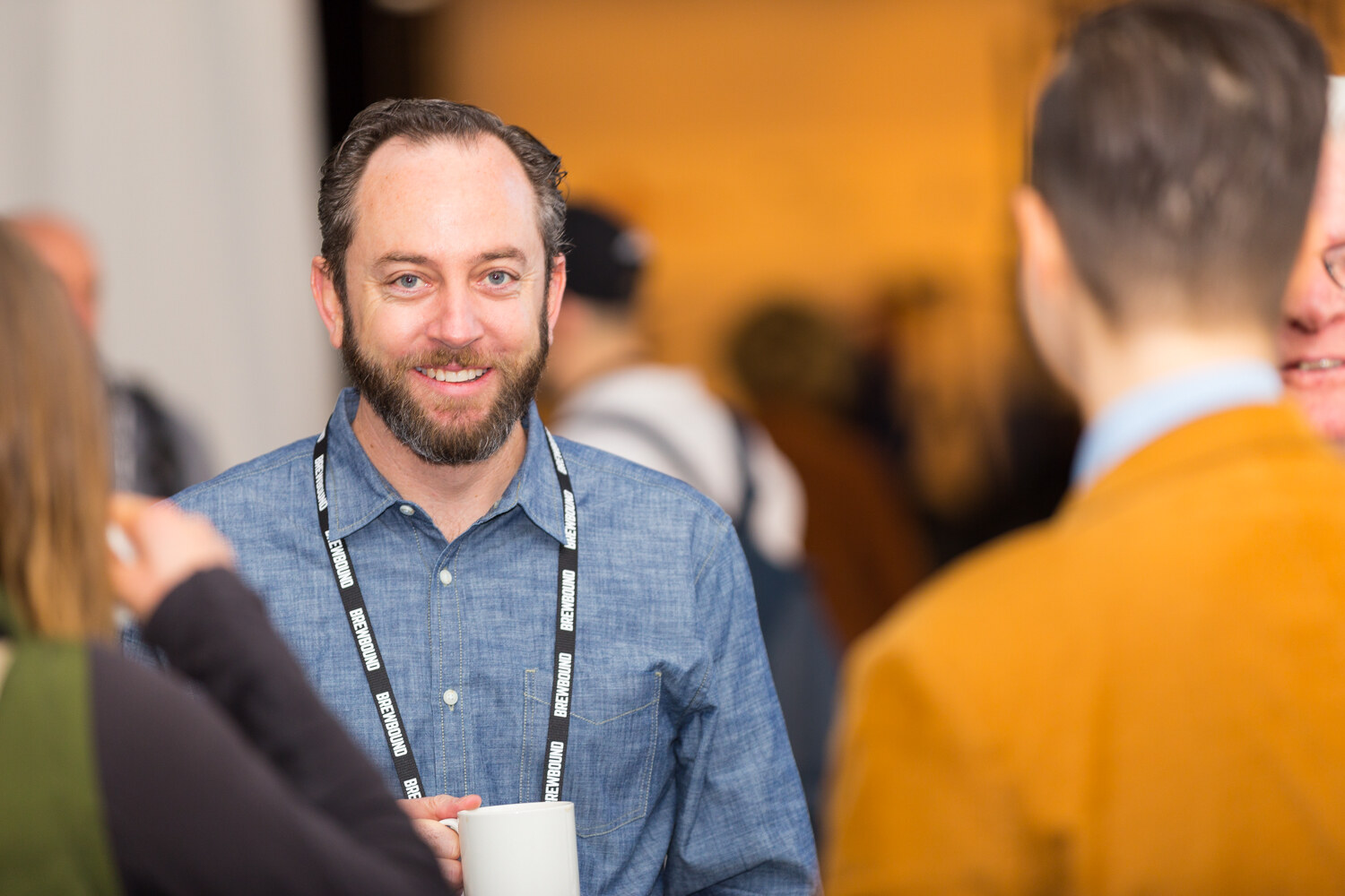 Man with a beard in a blue chambray shirt and a Brewbound lanyard holding a white mug, with warm blurred exhibit lights behind him.