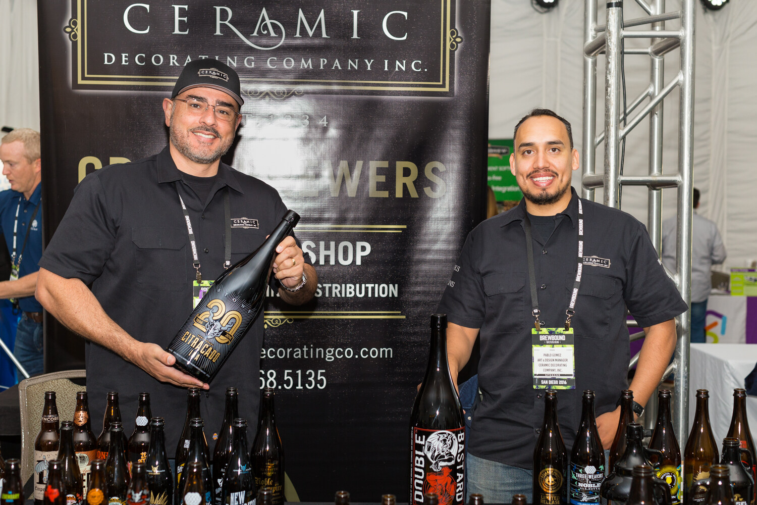 Two men in black work shirts and Brewbound Session badges smiling in front of a Ceramic Decorating Company Inc. banner, one holding a large black bottle, with decorated bottles in front.
