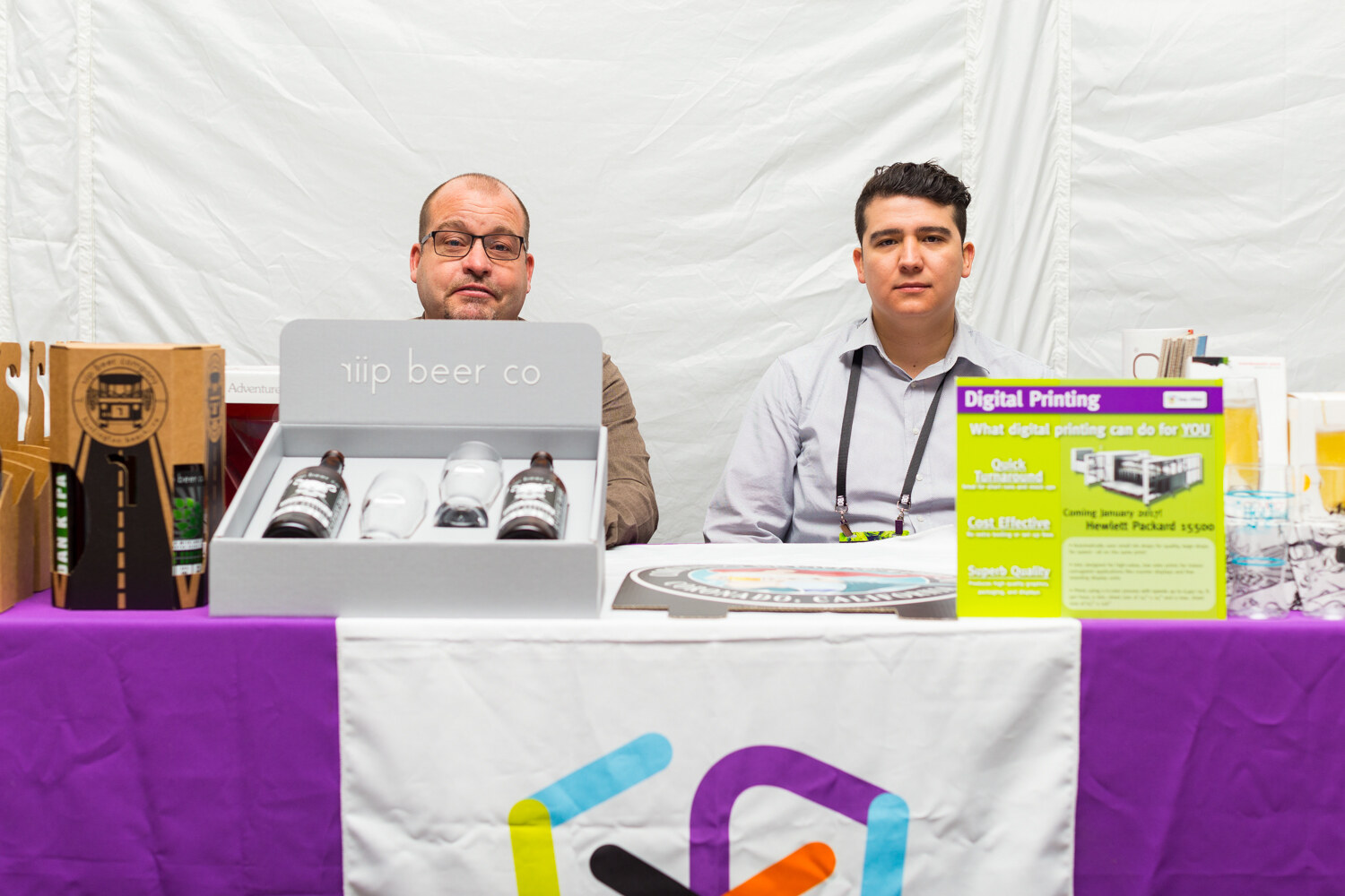Two seated men behind a purple-draped table holding a gray riip beer co box with two bottles and glasses, a Digital Printing flyer and a San Francisco beer carton.