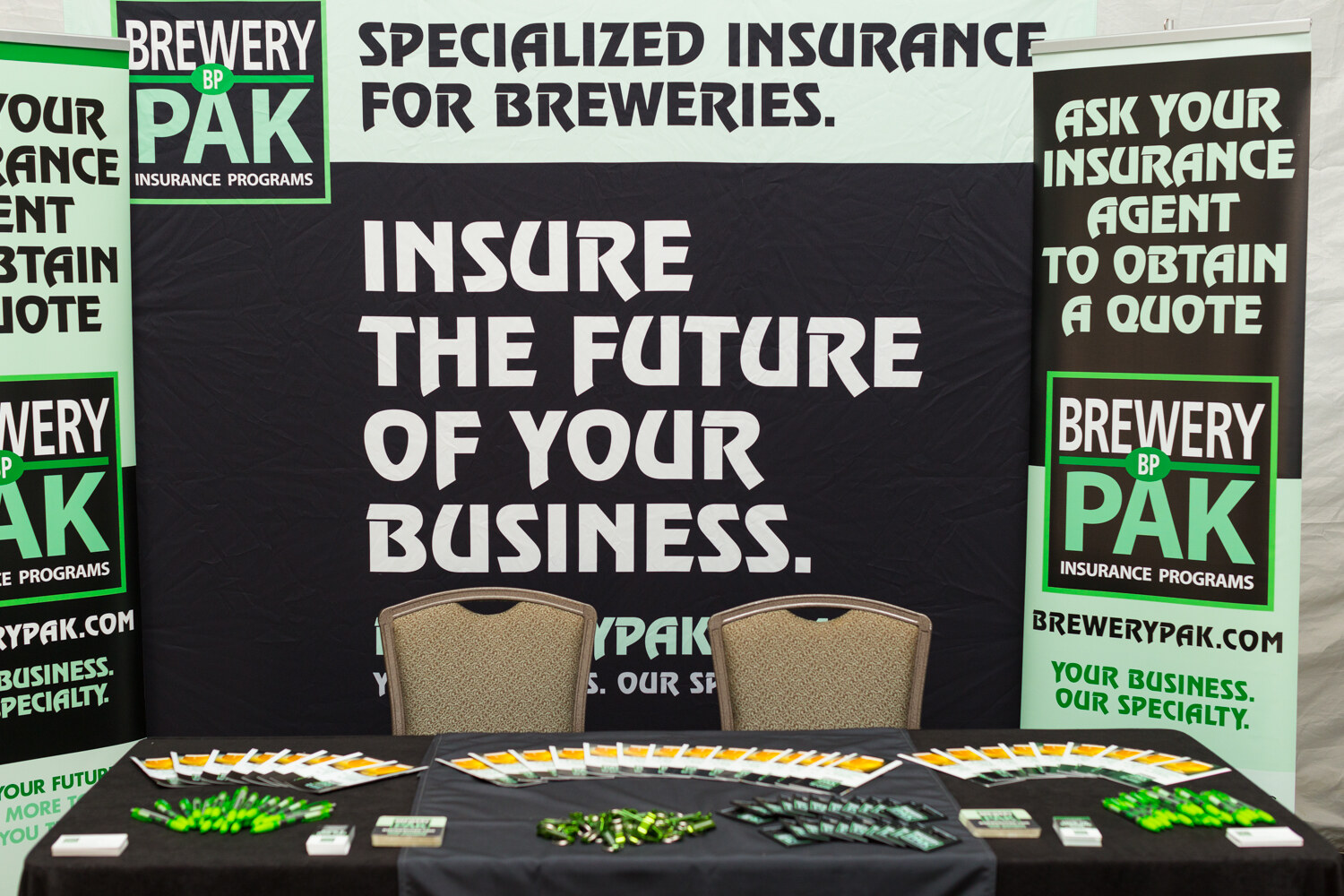 Black and white booth backdrop reading Specialized insurance for breweries, Insure the future of your business, with Brewery Pak banners and giveaways on a black table.