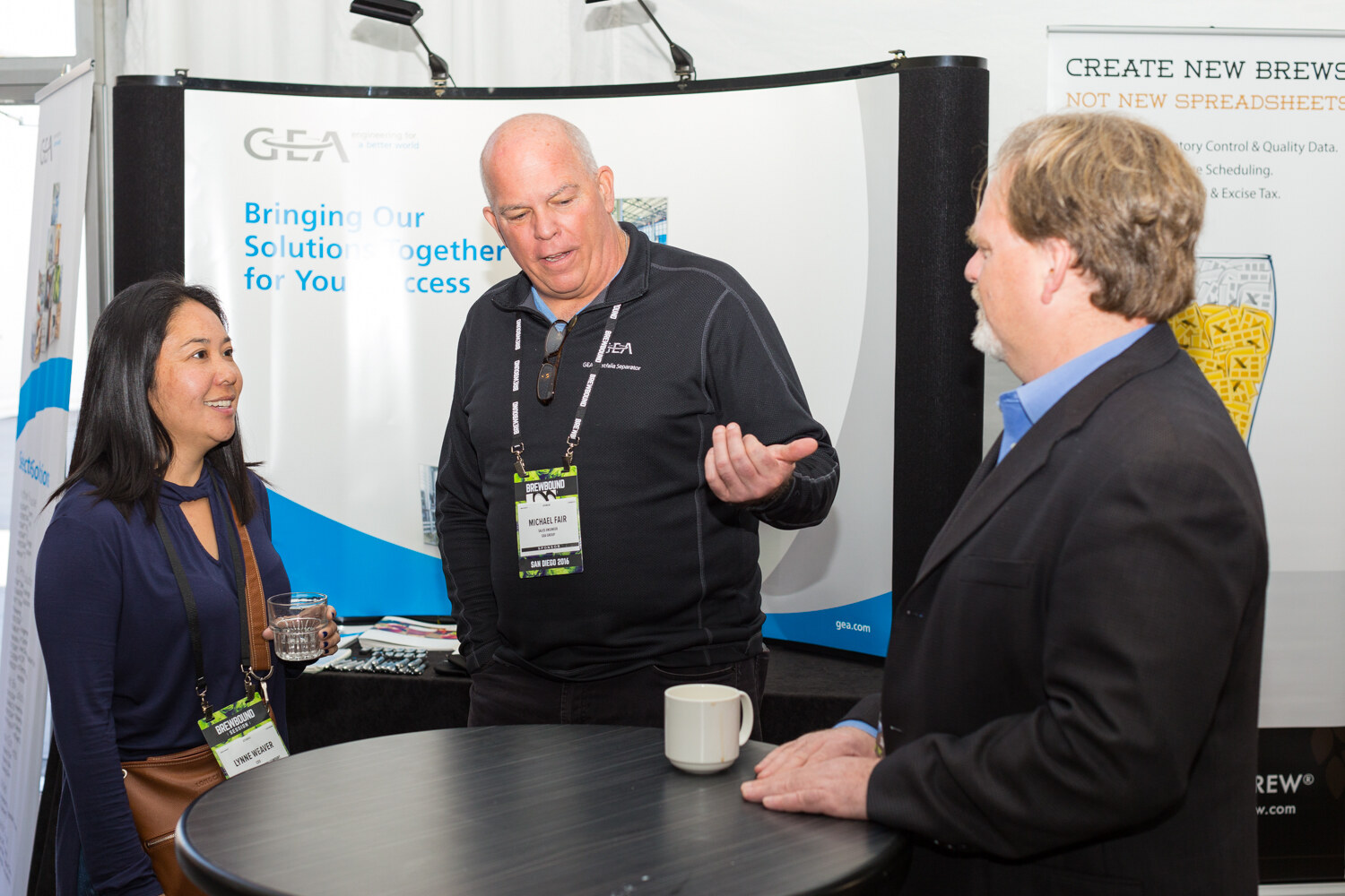Woman holding a glass of water, a man in a black GEA quarter-zip with a Brewbound badge and a man in a dark jacket gathered around a high table in front of a GEA banner reading Bringing Our Solutions Together for Your Success.