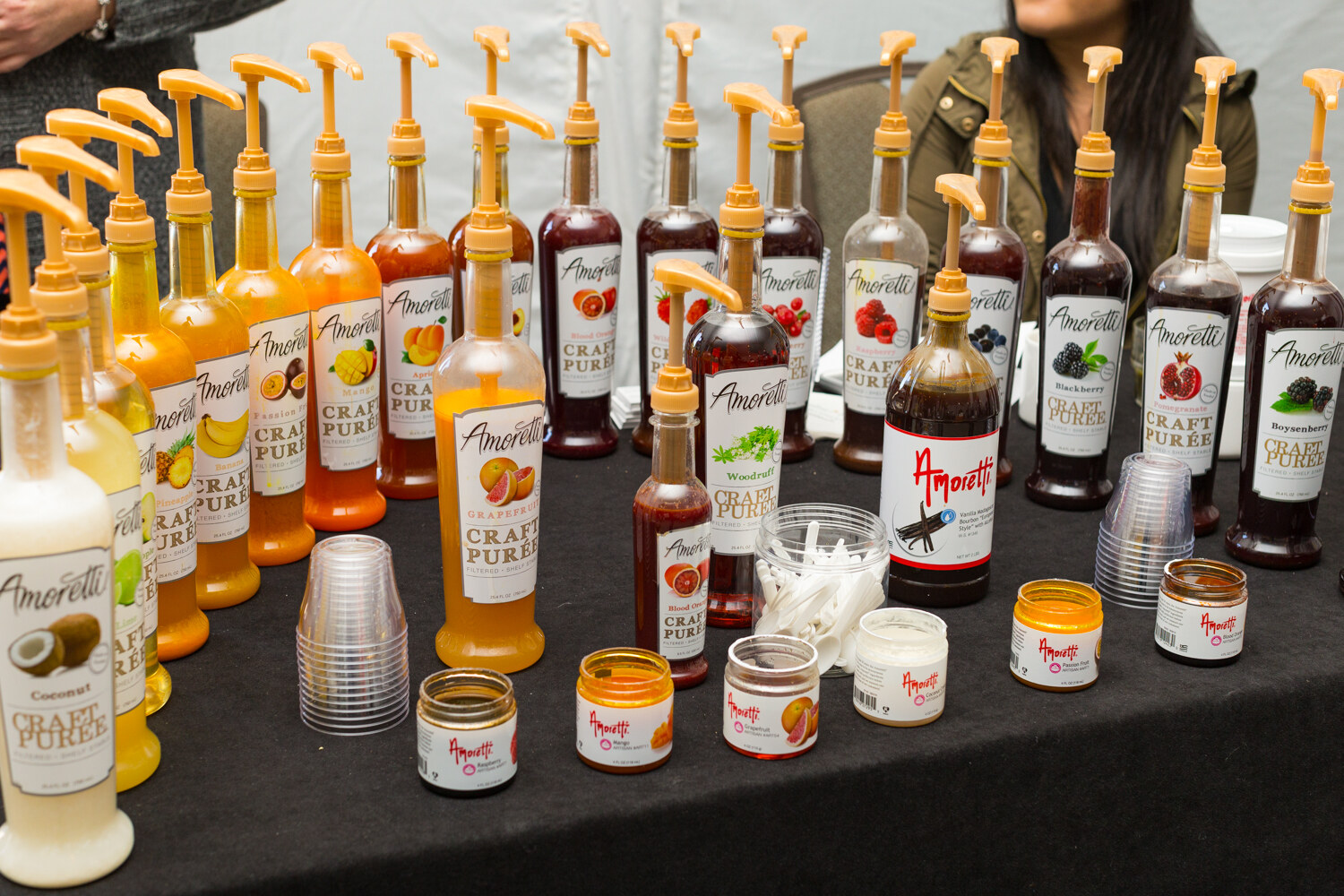 Dozens of Amoretti Craft Puree pump bottles in fruit flavors with small sample jars and stacks of plastic cups on a black table.