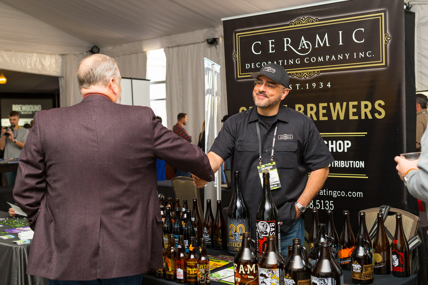 Man seen from behind in a burgundy blazer shaking hands with a smiling exhibitor in a black work shirt and cap in front of a Ceramic Decorating Company Inc. banner.