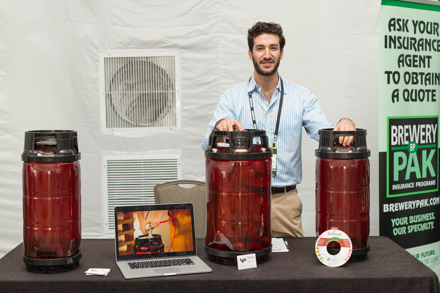 Smiling man in a light blue checked shirt with a lanyard resting his hands on two red plastic kegs, with a laptop and a Brewery Pak banner on the table.