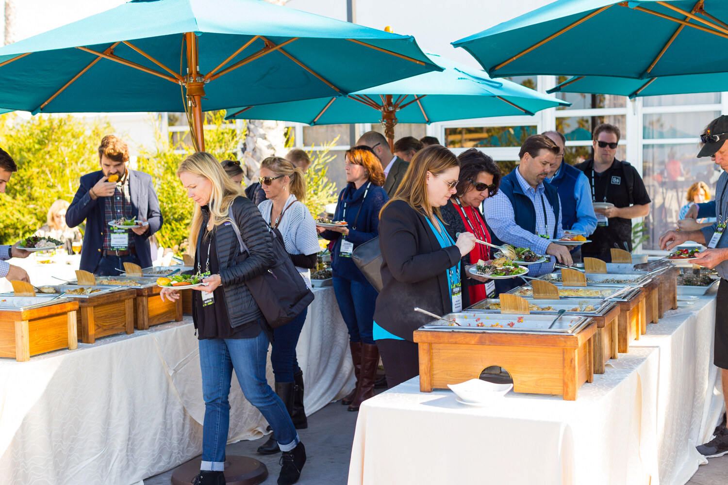 Attendees with lanyards serving themselves at chafing dishes on white-clothed tables under teal patio umbrellas in sunshine.