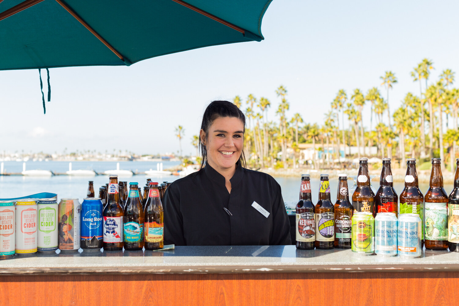 Woman in a black jacket behind a bar holding up a row of beer cans and bottles including Long Board, Coronado and Pabst, with a lagoon and palm trees in the background.