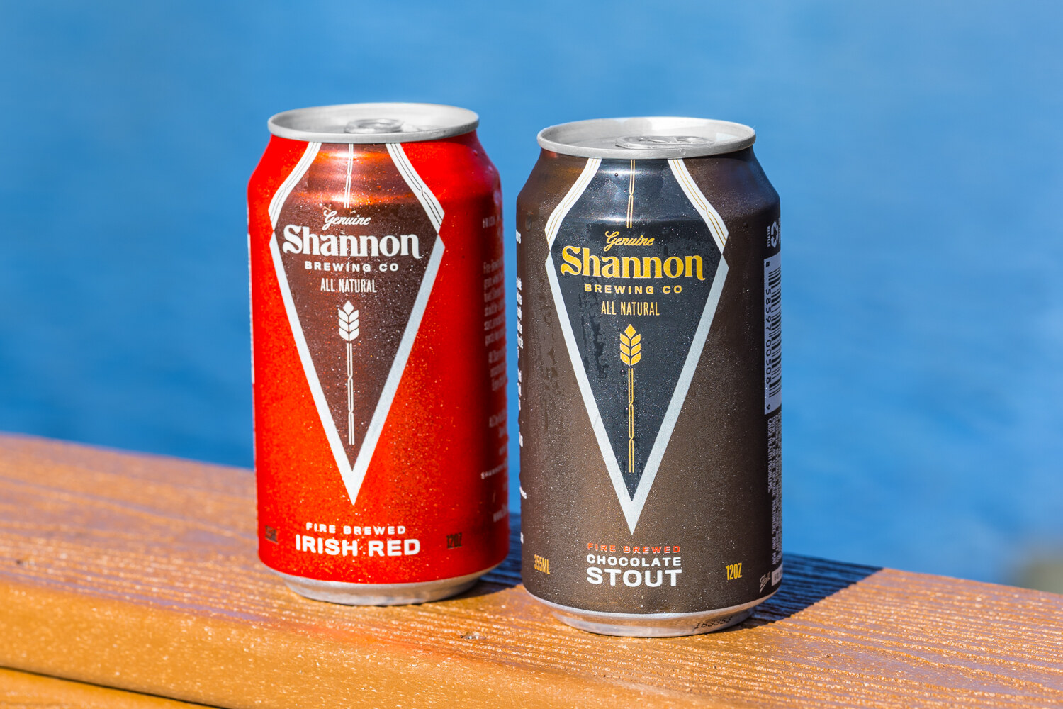 Two cans labeled Genuine Shannon Brewing Co, one red Irish Red and one dark Chocolate Stout, on a wooden railing with blue water behind.