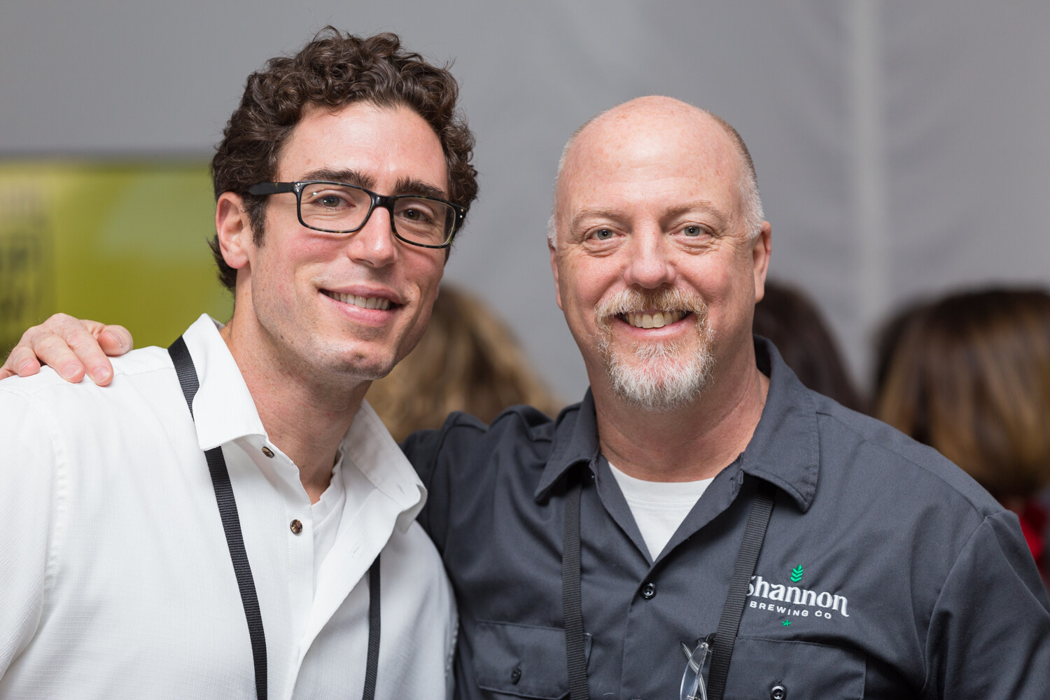 Curly-haired man in glasses and a white shirt with his arm around a bald man in a gray Shannon Brewing Co. work shirt, both smiling at the camera.