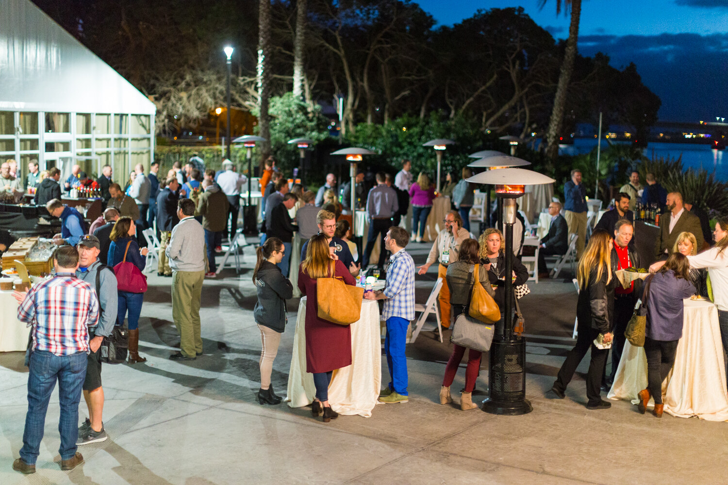High-angle view of attendees standing around draped tables and patio heaters beside a white tent at dusk, with the bay in the distance.