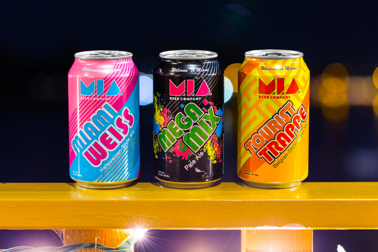 Three cans labeled M.I.A. Beer Company: a pink and blue Miami Weiss, a black Mega Mix Pale Ale and an orange Tourist Trappe, against a dark background with blurred lights.