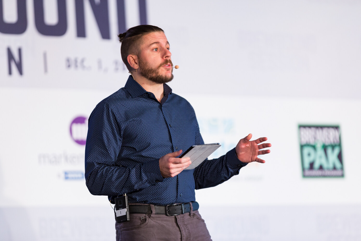 Bearded man with a headset microphone and a navy patterned shirt holding a tablet, in front of a banner reading Dec. 1 with Markem-Imaje and Brewery Pak logos.