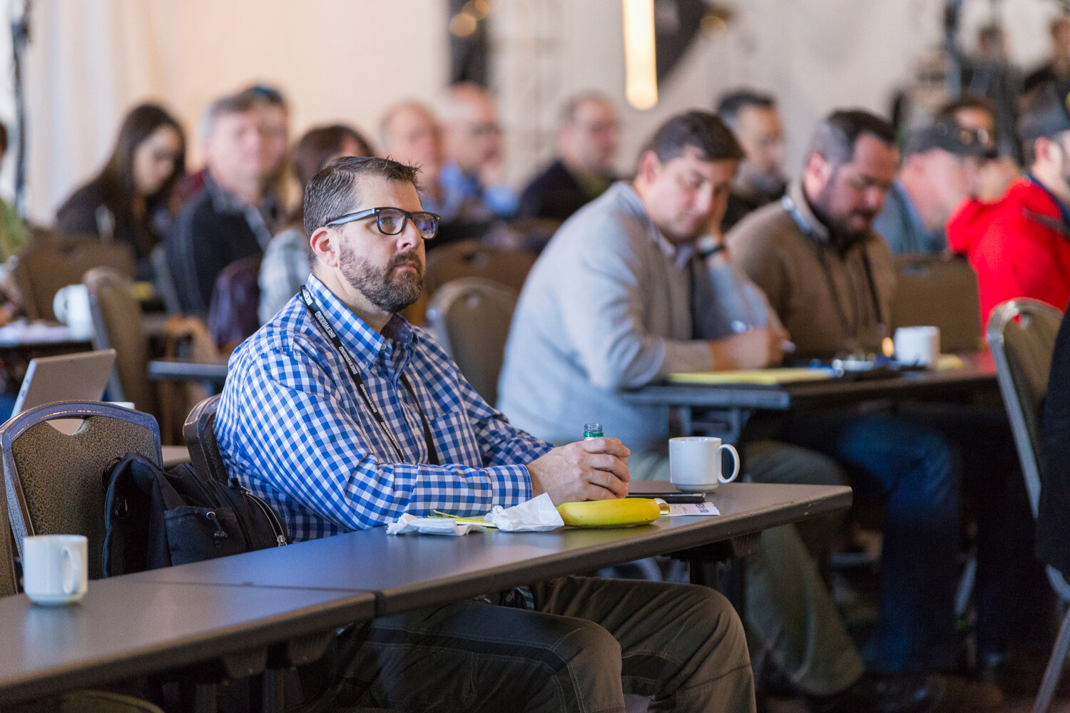 Bearded man in glasses and a blue checked shirt at a long table with a coffee cup and a banana, with rows of attendees blurred behind him.